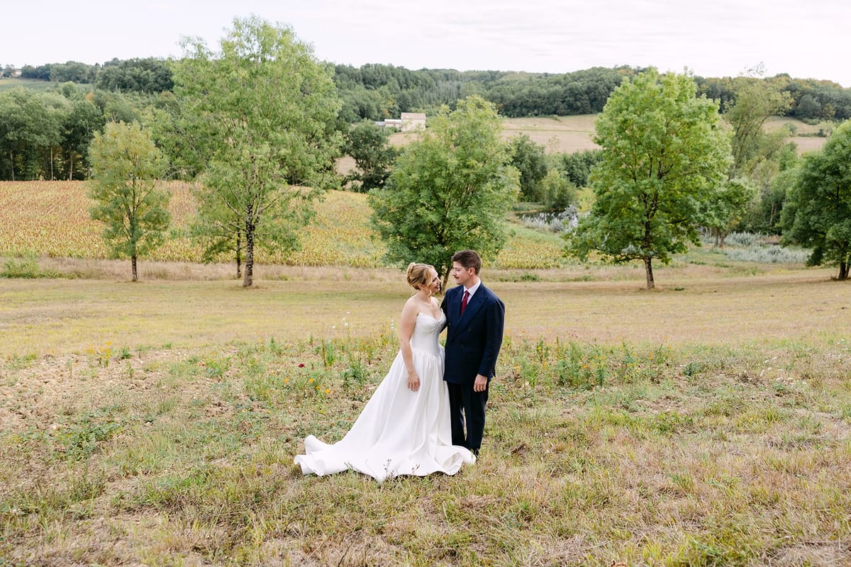 Bride and groom stand in a meadow among trees, with fields and wooded hills rolling behind