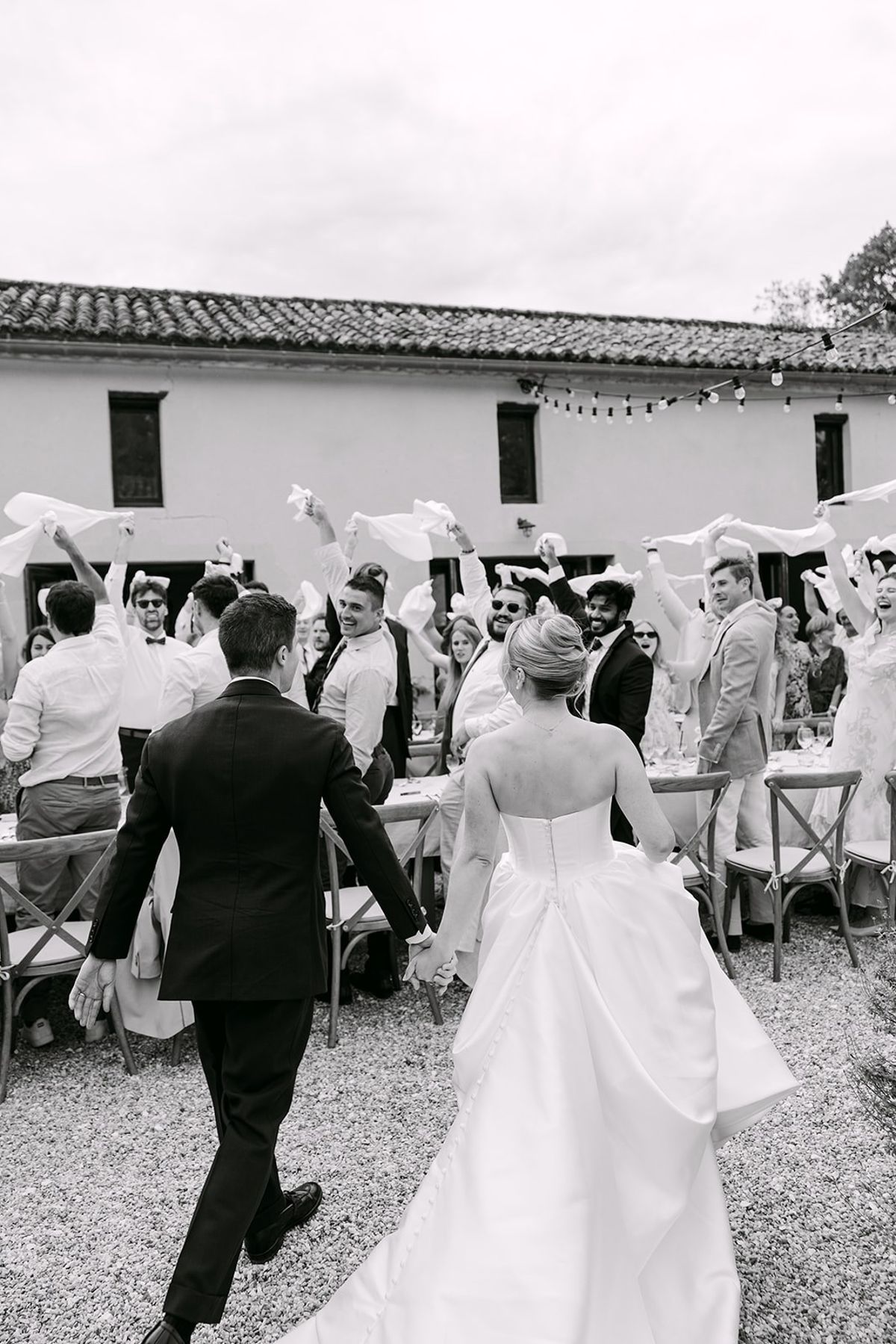 Bride and groom walk hand in hand towards guests waving white napkins at long tables on gravel, in black and white