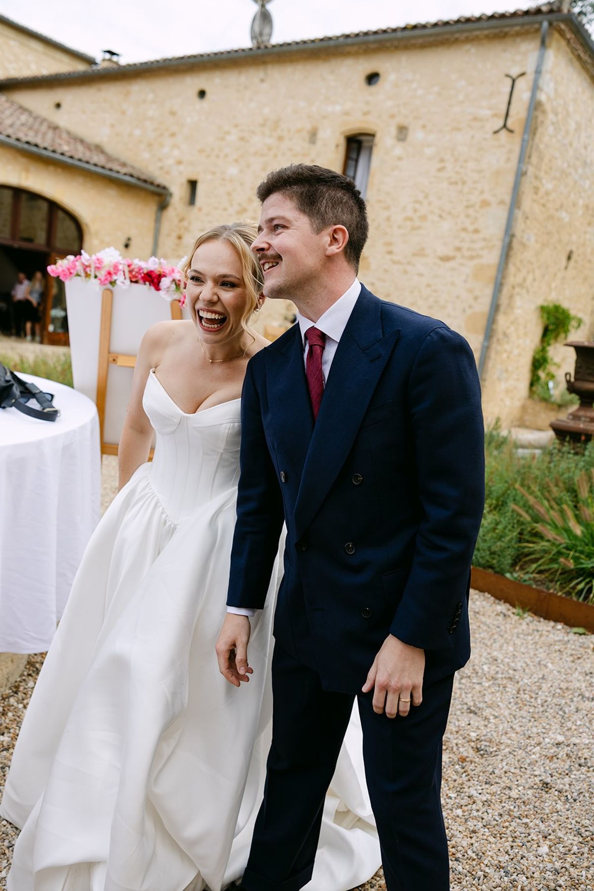 Bride and groom laugh on the gravel courtyard in front of the stone château with its arched doorway