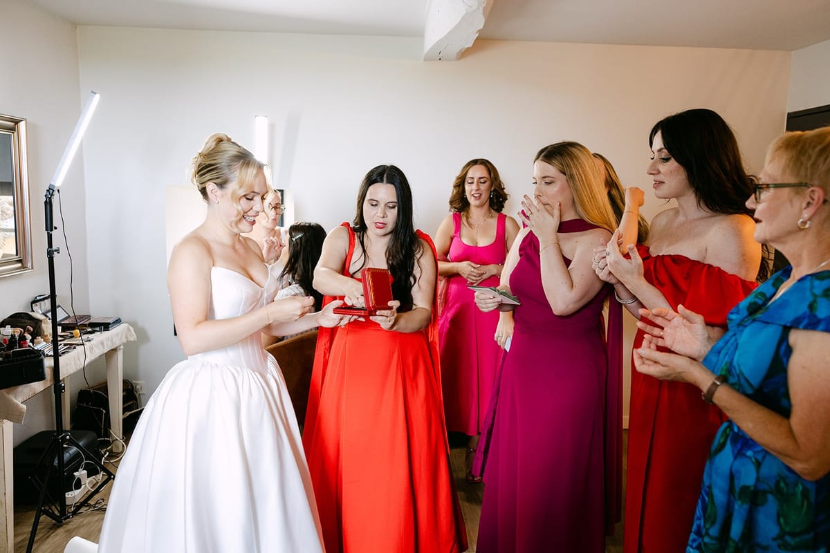 Bride in a strapless gown takes something from a red box held by a woman in red, other women in pink and red nearby