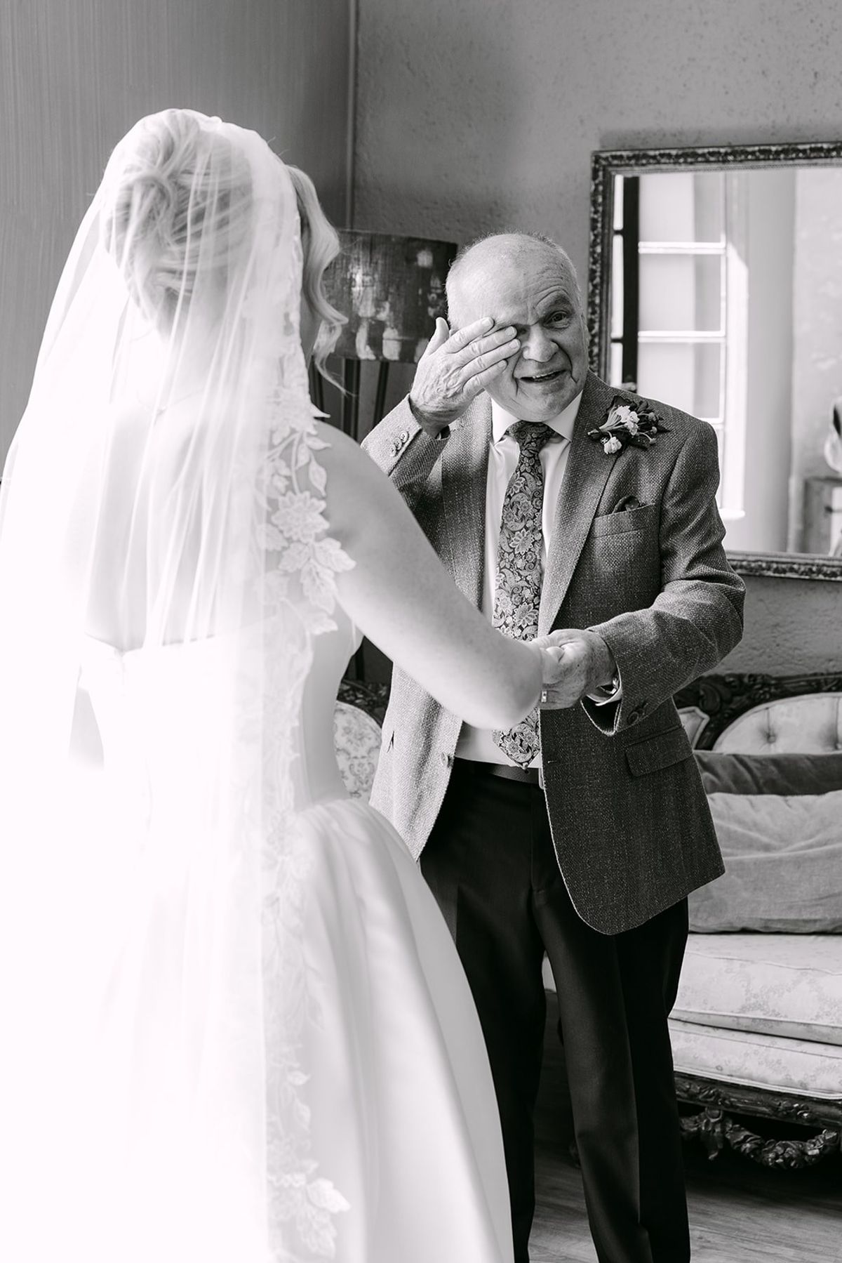 Older man wipes his eye while holding the bride's hand in a room with an ornate mirror and sofa, in black and white