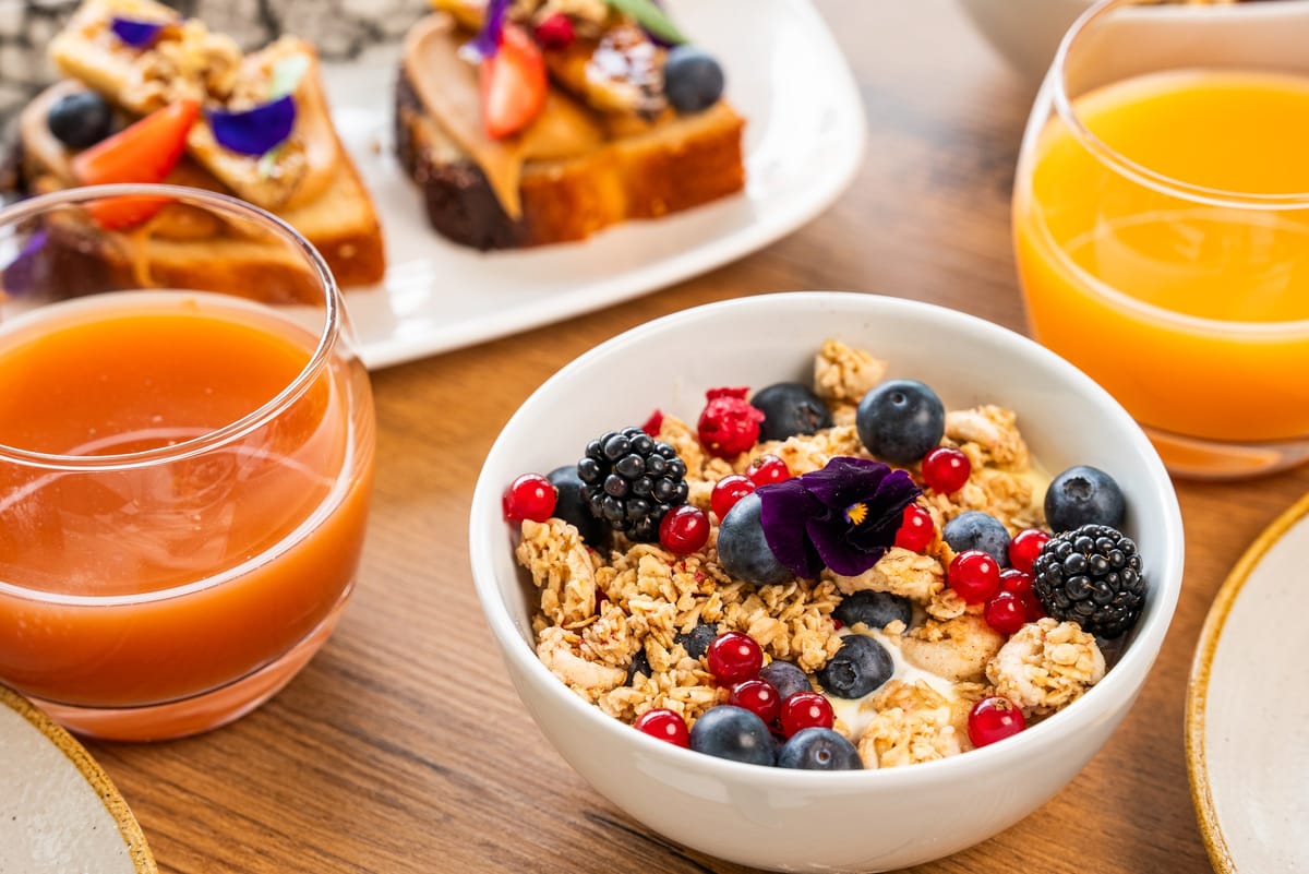 Bowl of granola with blackberries, blueberries, redcurrants and a pansy between two glasses of juice