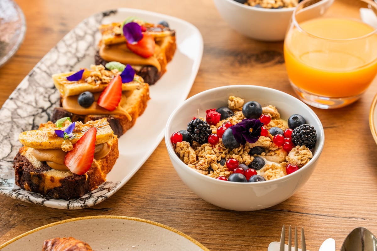 Granola bowl with fresh berries beside a platter of banana-topped cake slices and a glass of orange juice