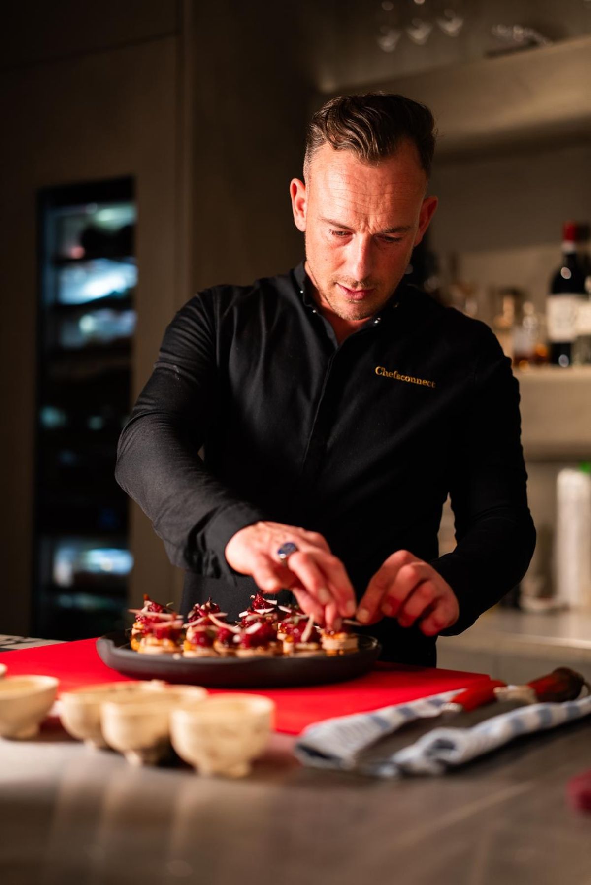 Chef in a black jacket arranging canapés with red topping on a dark plate on a kitchen counter