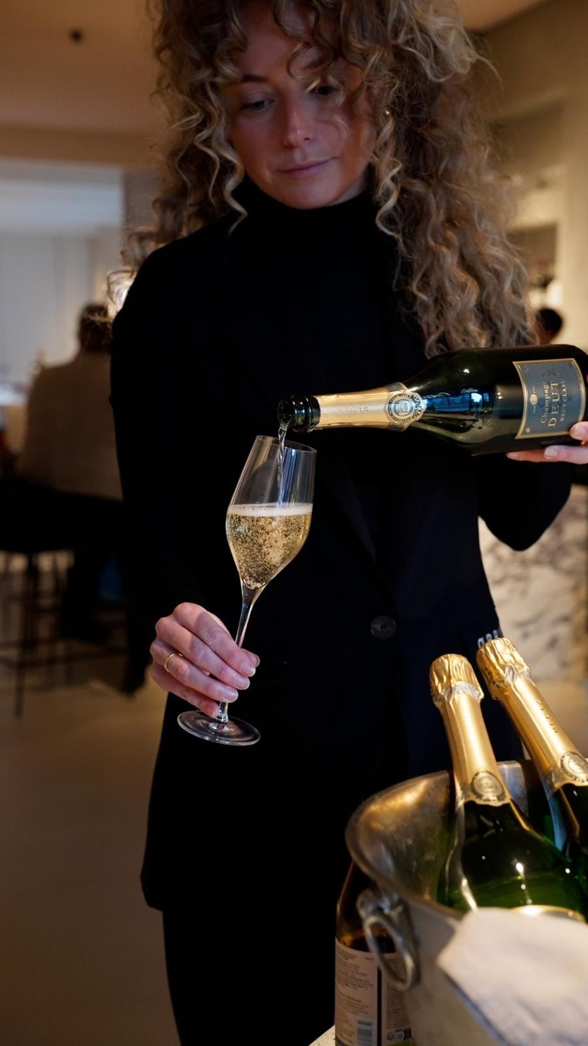 Woman in black pours champagne into a flute beside an ice bucket of bottles