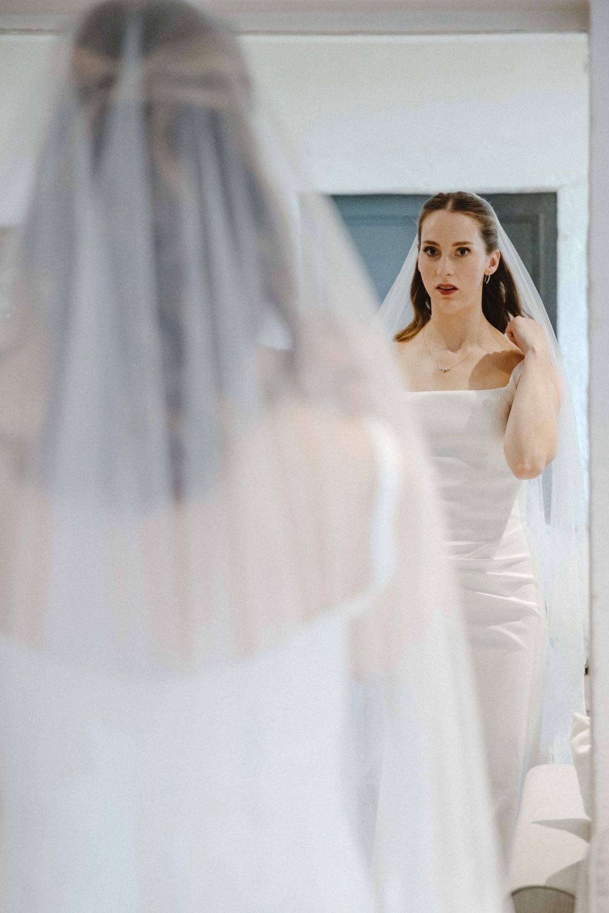 Bride in a square-neck gown and veil seen in a mirror in a white room with a grey-blue door