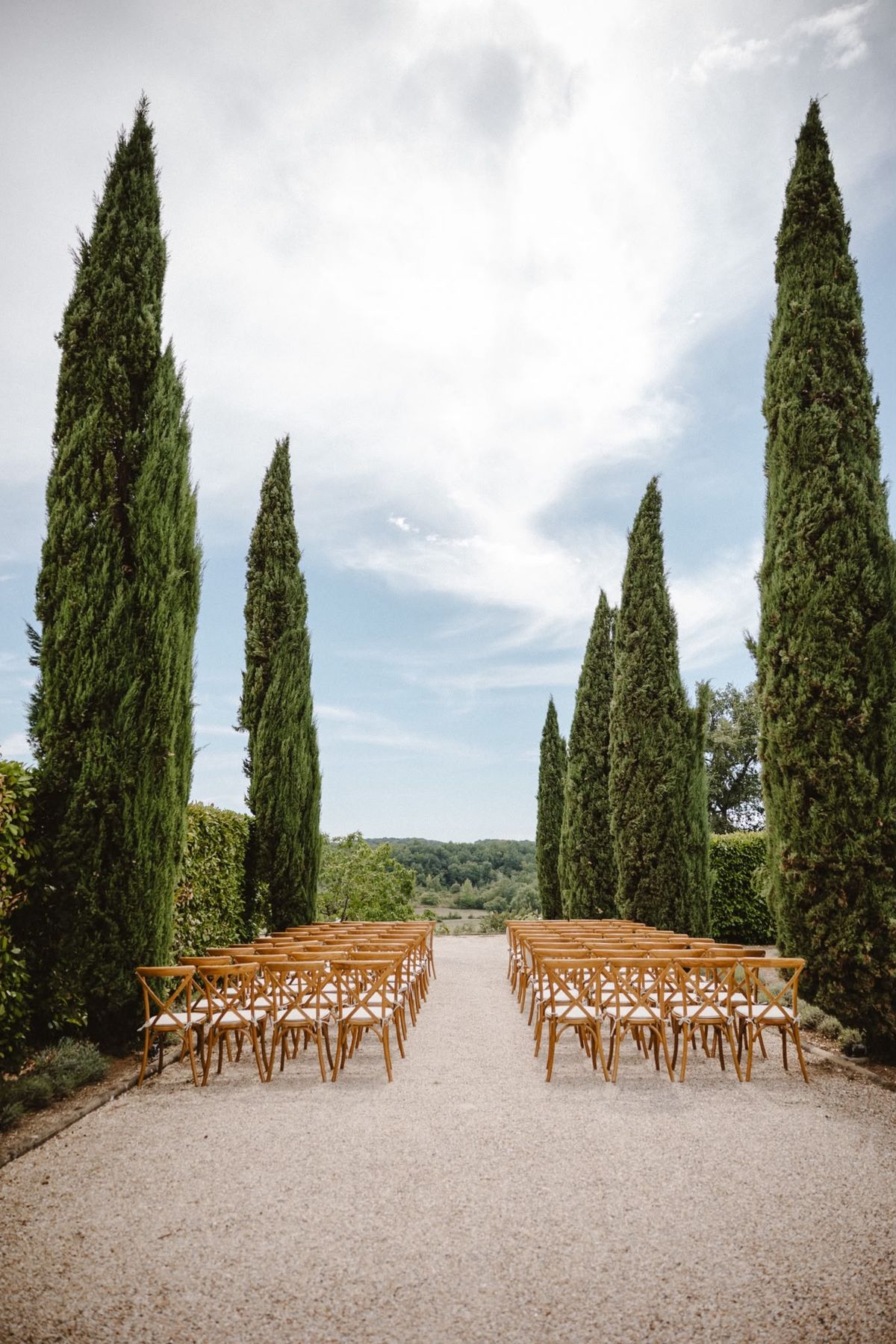 Rows of wooden cross-back chairs on the gravel cypress avenue, framing a view of wooded hills
