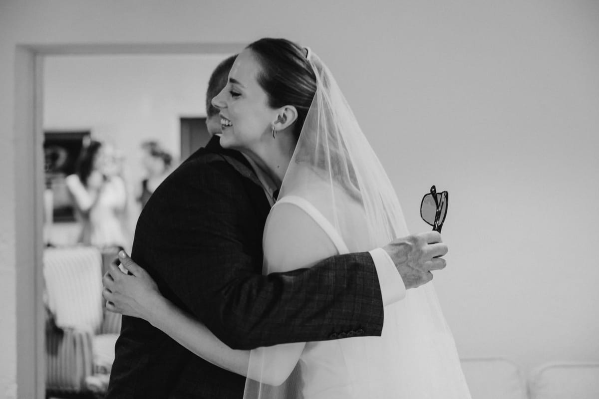 Bride in a veil hugs a man in a checked jacket, guests visible through a doorway behind, in black and white