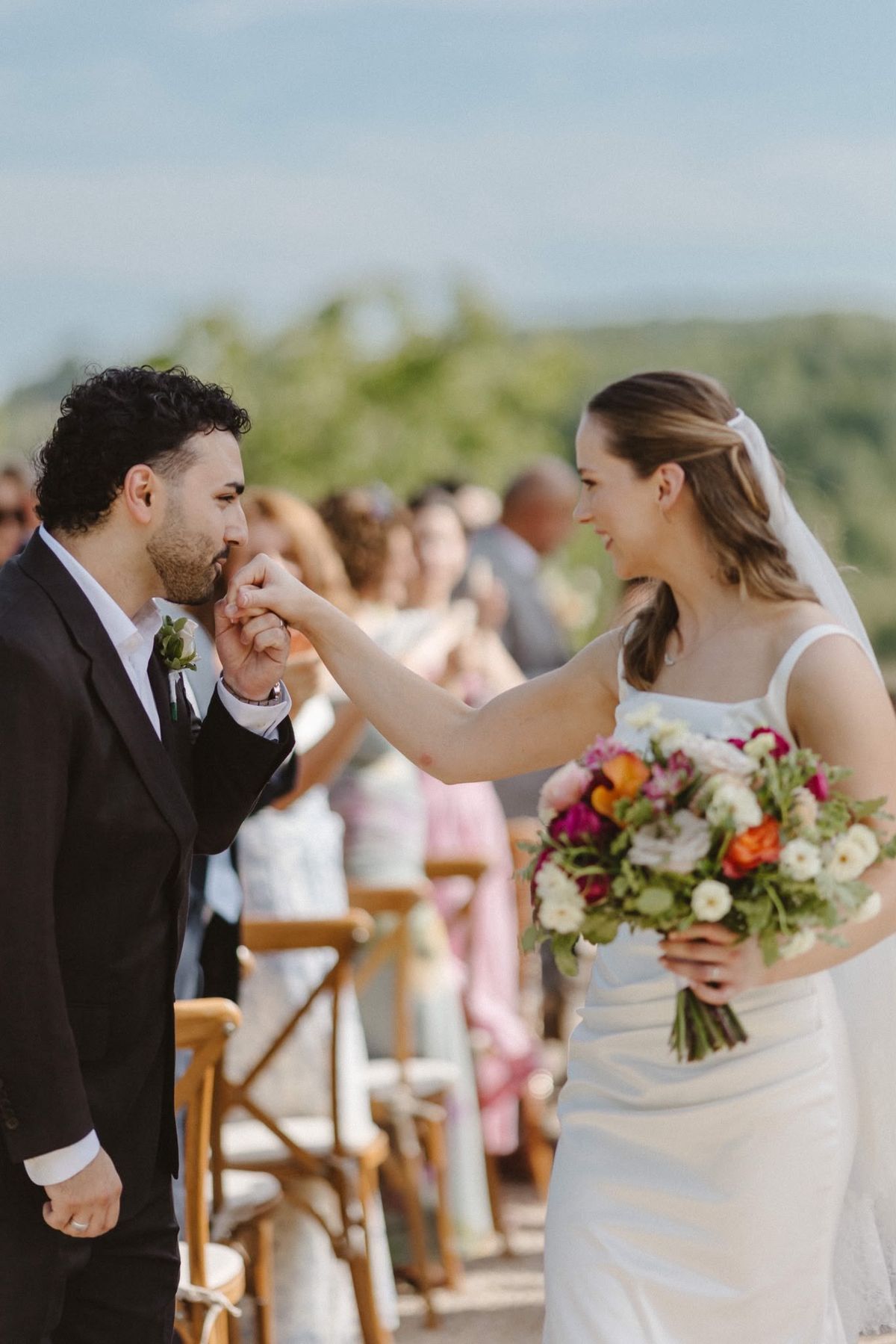 Groom kisses the bride's hand in the aisle between cross-back chairs and guests, hills blurred behind