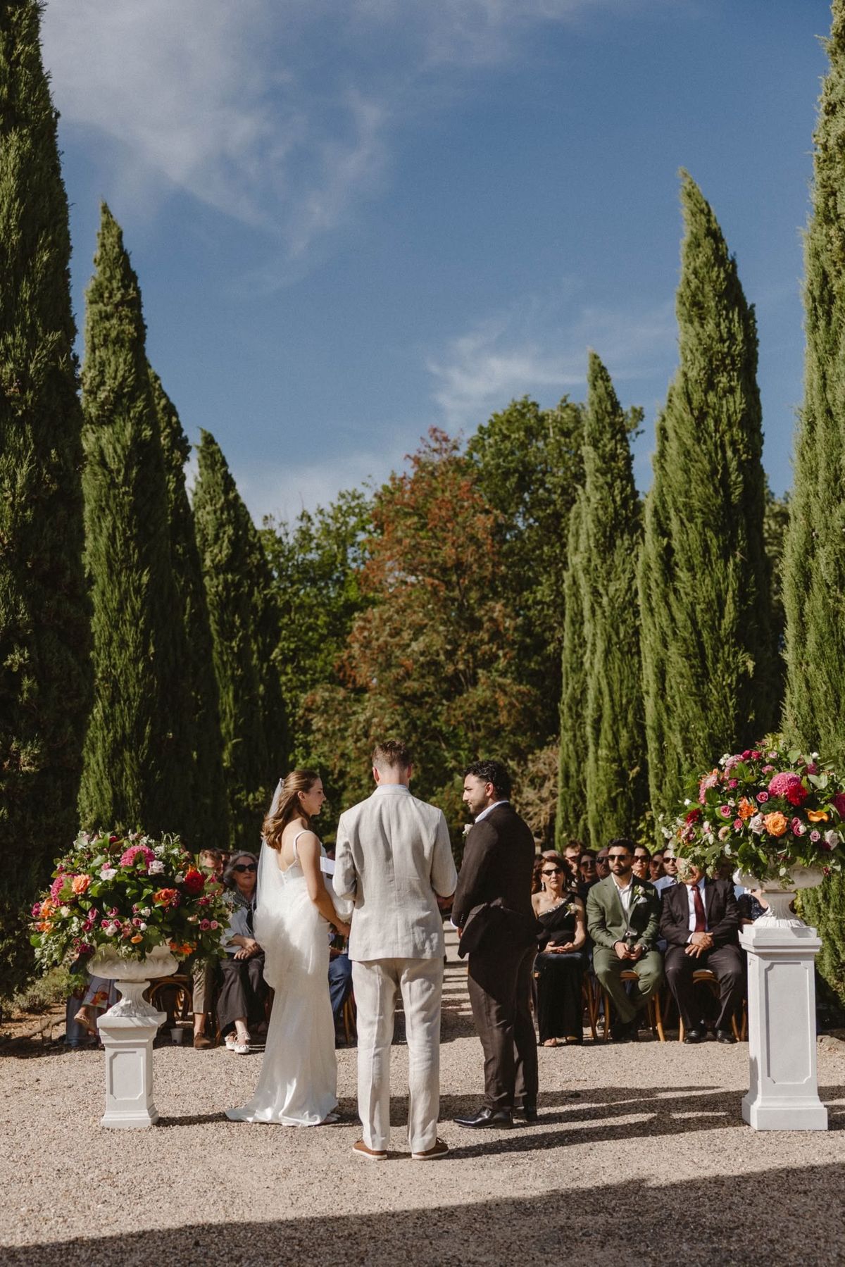 Bride and groom stand before a man in a pale suit on the gravel cypress avenue, flower urns either side and guests seated
