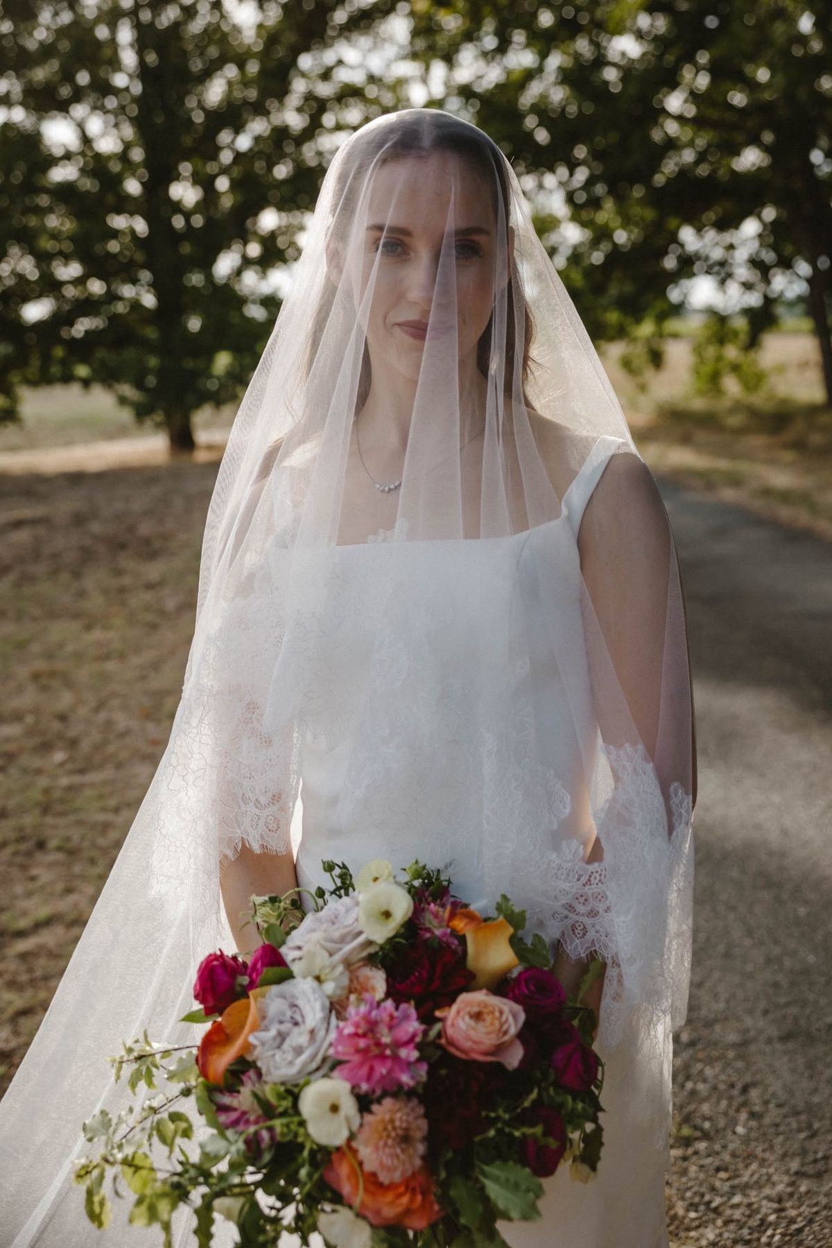 Bride with her veil over her face holding a pink, orange and red bouquet on a tree-lined path