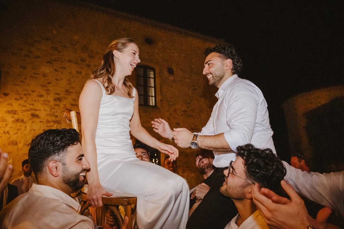 Bride and groom lifted on chairs by laughing guests at night in front of the warmly lit stone château