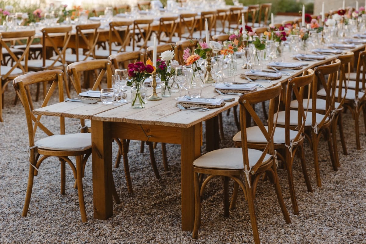 Long wooden tables with cross-back chairs on gravel, set with candles, bud vases and woven placemats