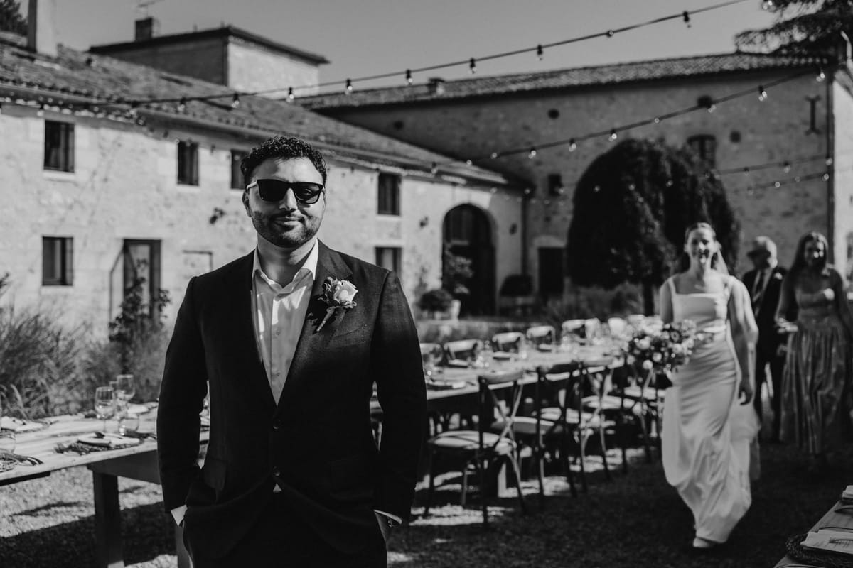 Groom in sunglasses waits as the bride walks up behind him past long tables and festoon lights, in black and white