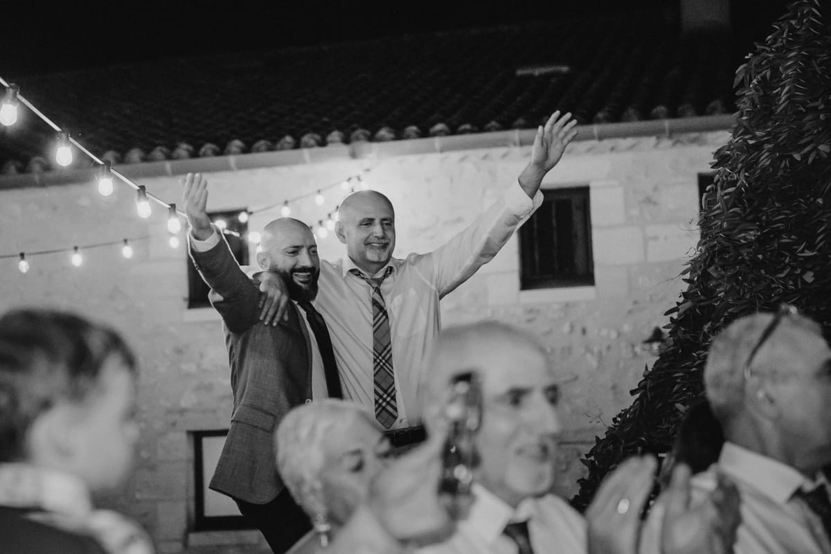 Two men raise their arms above the crowd at night under festoon lights by a stone building, in black and white