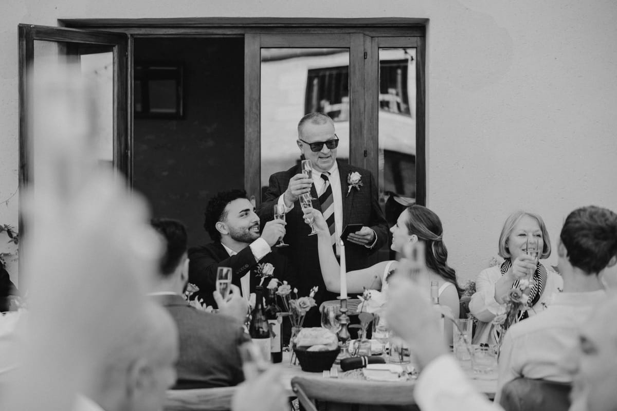 Man in sunglasses raises a glass to the bride and groom at a candlelit table by glazed doors, in black and white