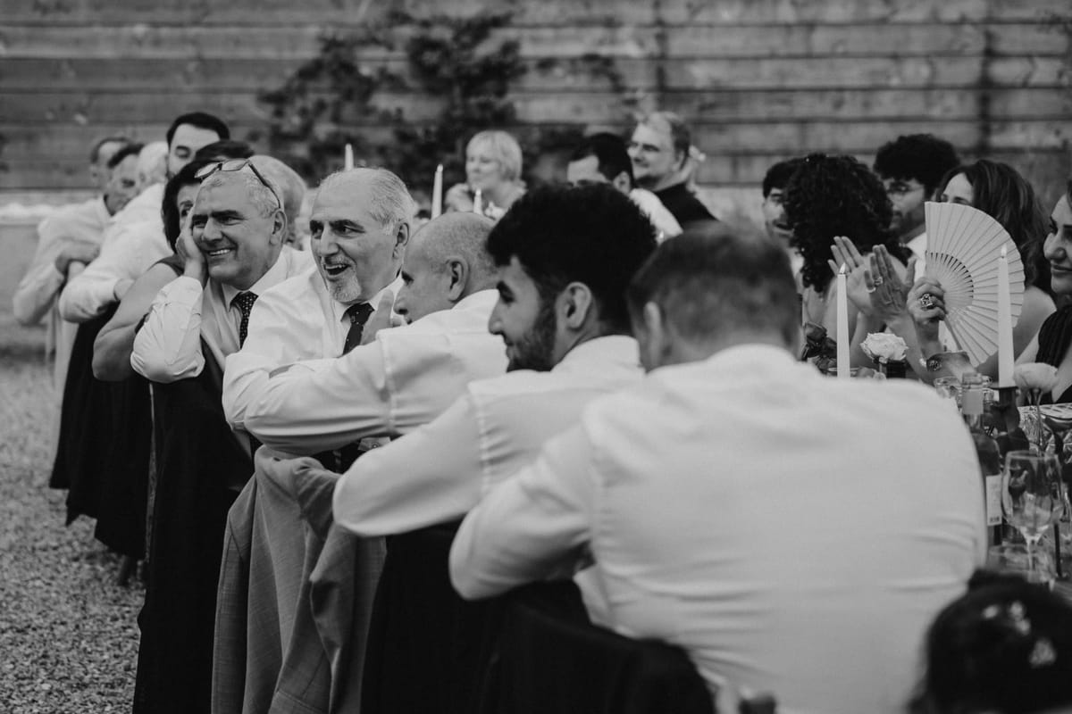 Guests laugh and clap along a candlelit table on gravel, one woman holding a fan, in black and white