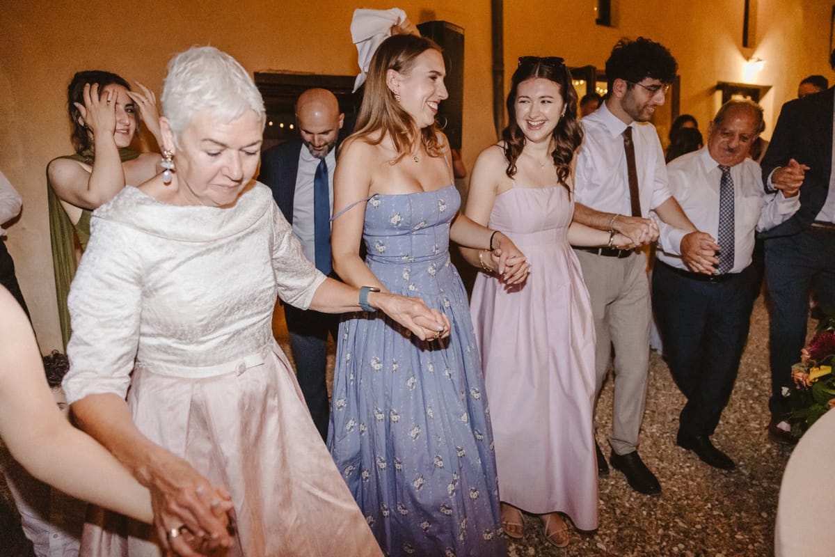 Guests dance hand in hand in a line on gravel at night beside a warmly lit rendered building