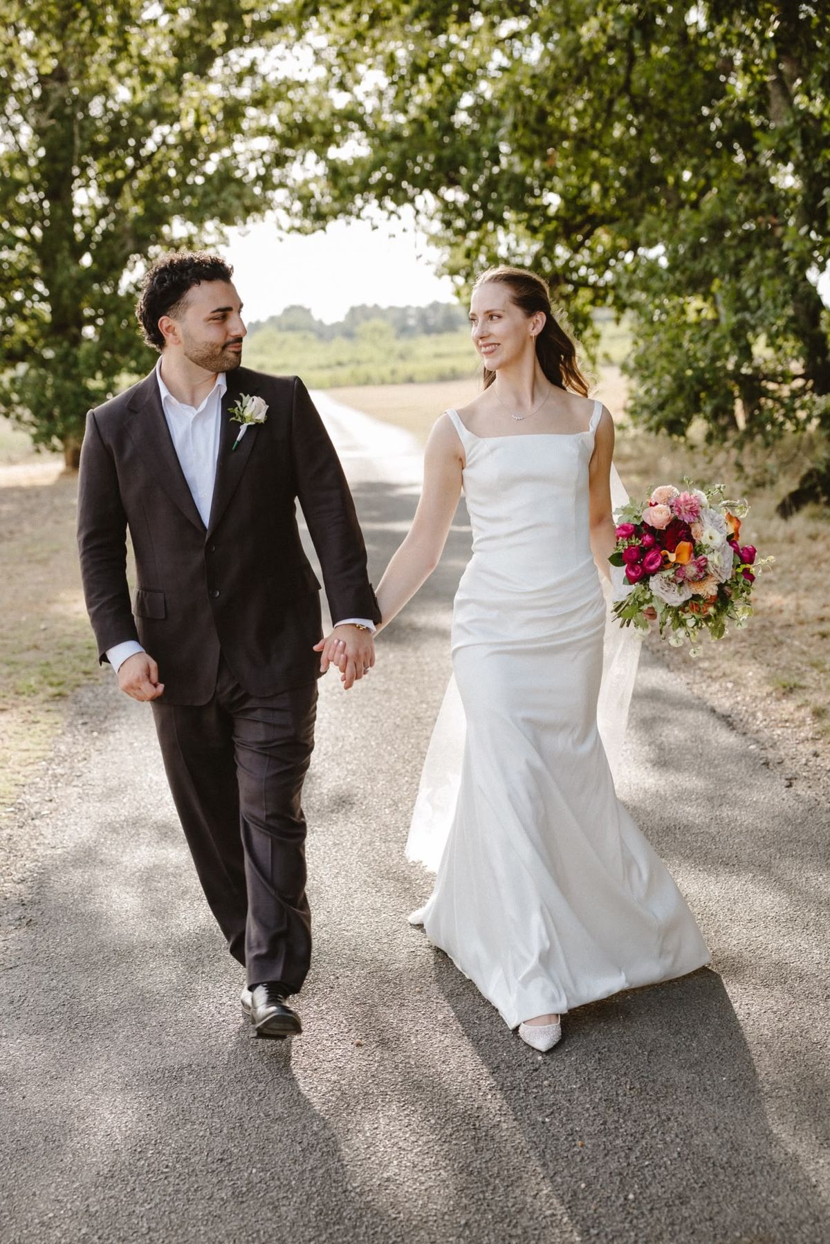 Bride and groom walk hand in hand down a tree-shaded drive, bride carrying a bright pink and orange bouquet