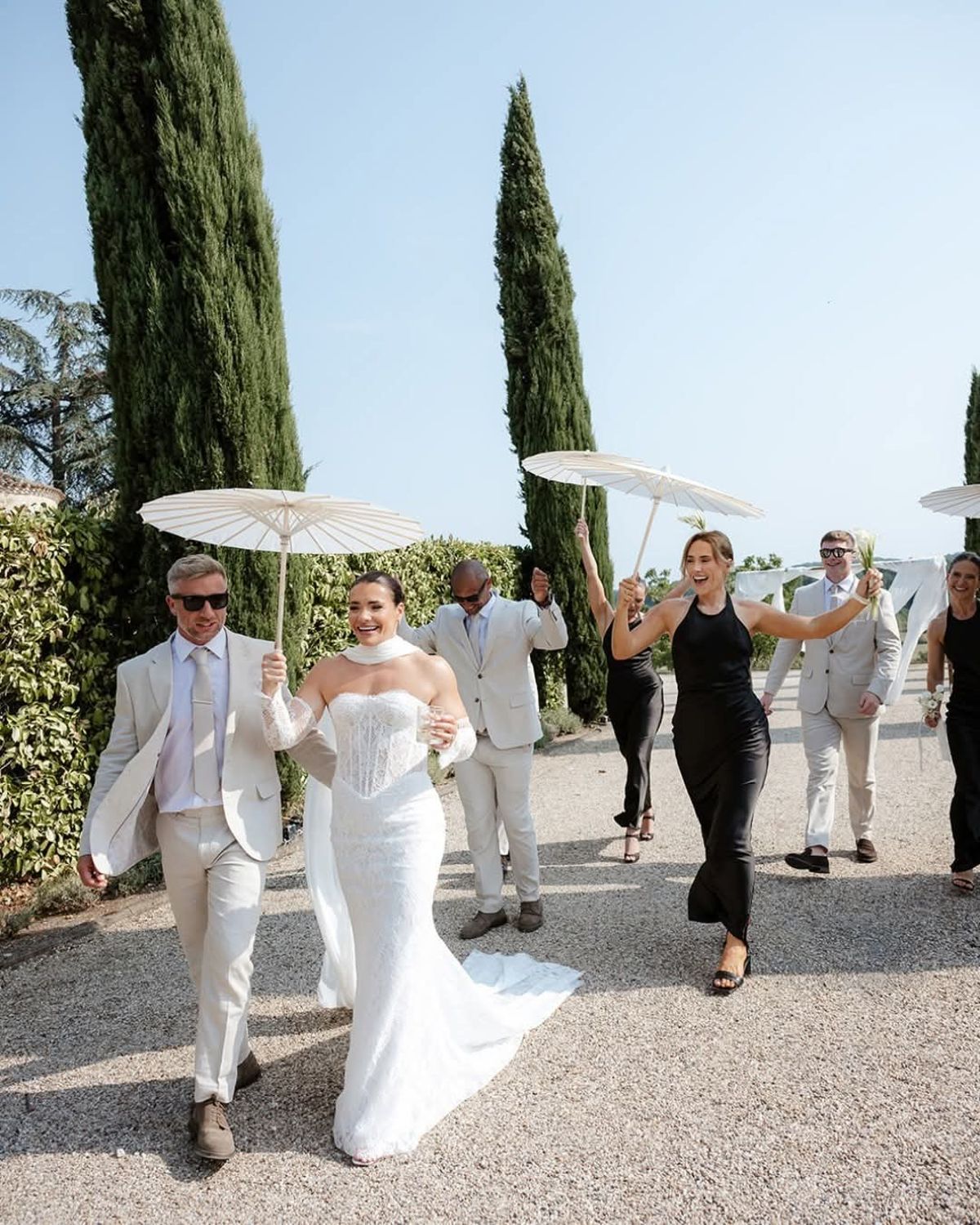 Bride walks with a man in a cream suit under a white parasol, guests following along the gravel cypress avenue
