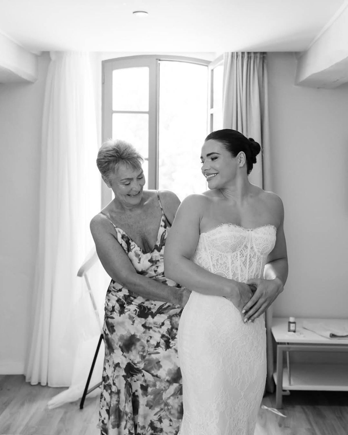 Woman in a floral dress adjusts the back of the bride's lace gown by a tall window, in black and white