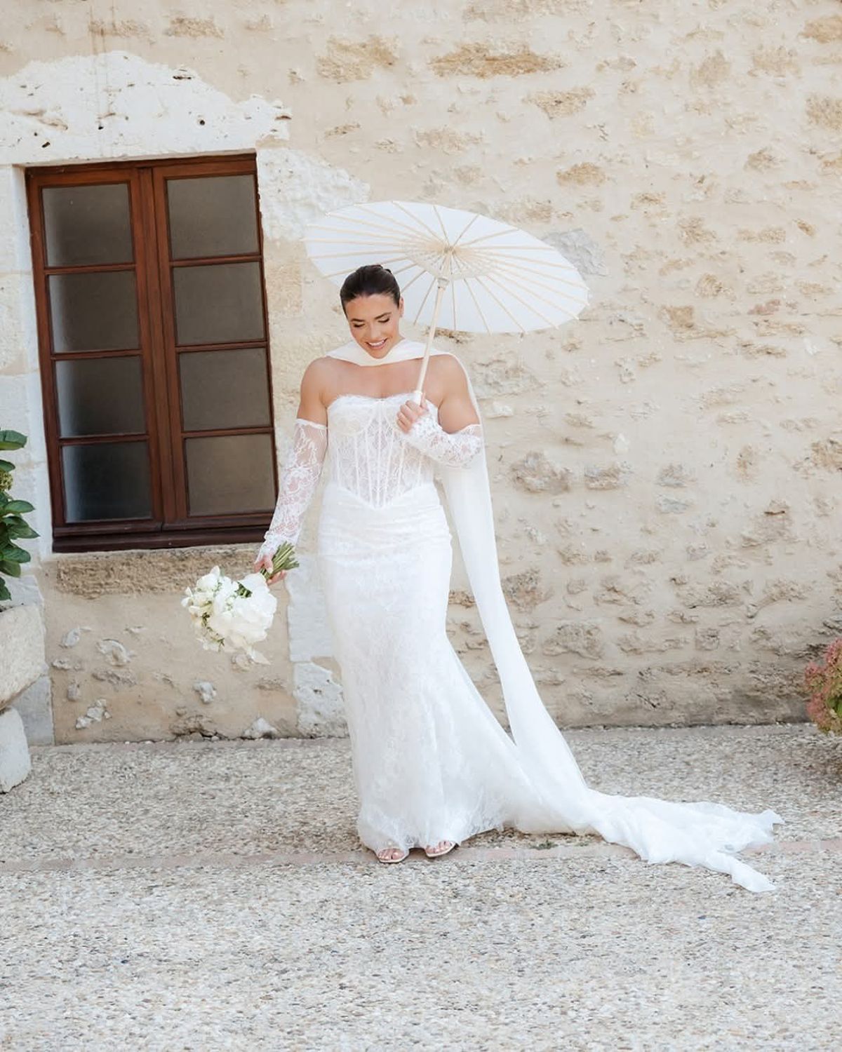 Bride in a lace gown and long scarf holds a white parasol and bouquet on gravel before a stone wall