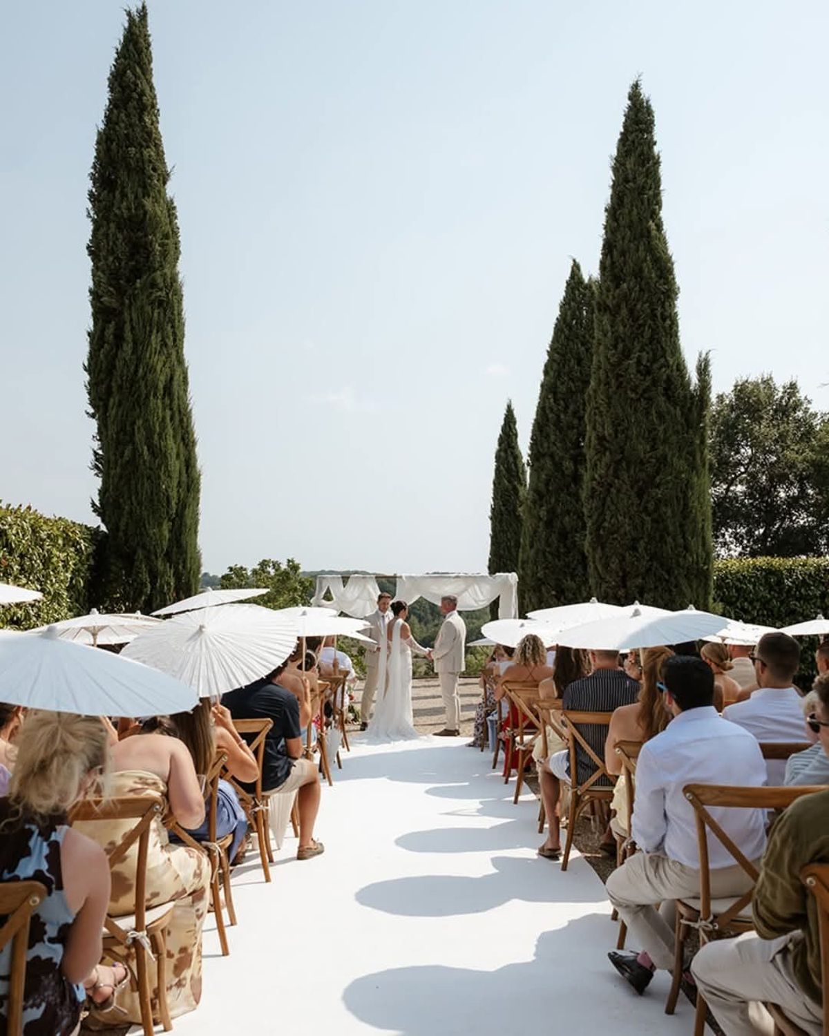 Guests with white parasols line a white aisle towards a draped arch at the end of the cypress avenue