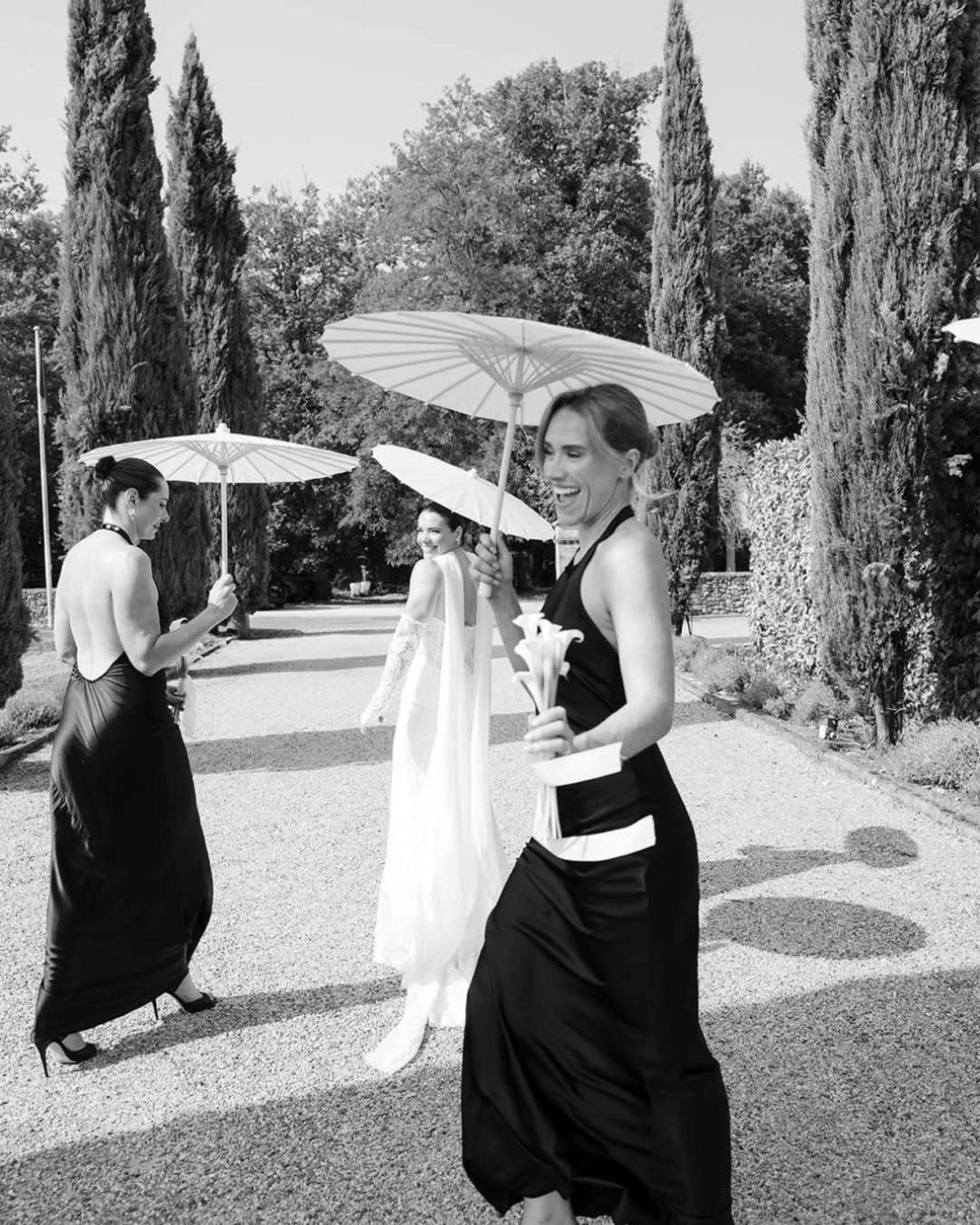 Bride looks back between two women in black dresses carrying parasols on the gravel cypress avenue, in black and white