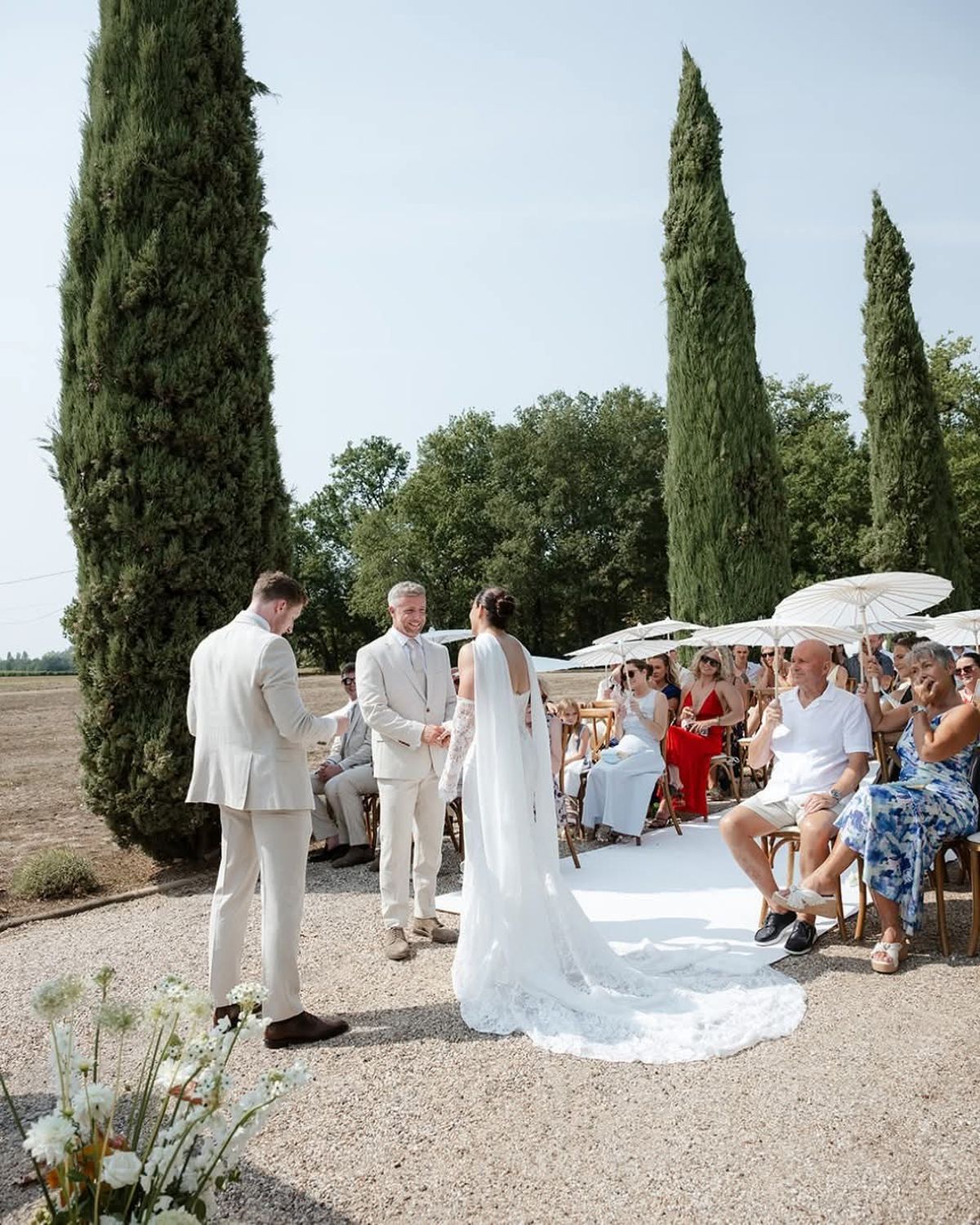 Bride faces a smiling man in a cream suit, a second man in cream beside them, guests with parasols seated by cypresses