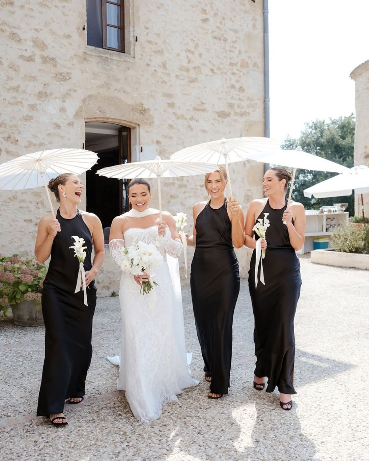 Bride and three women in black halter dresses carry white parasols across gravel before the stone château