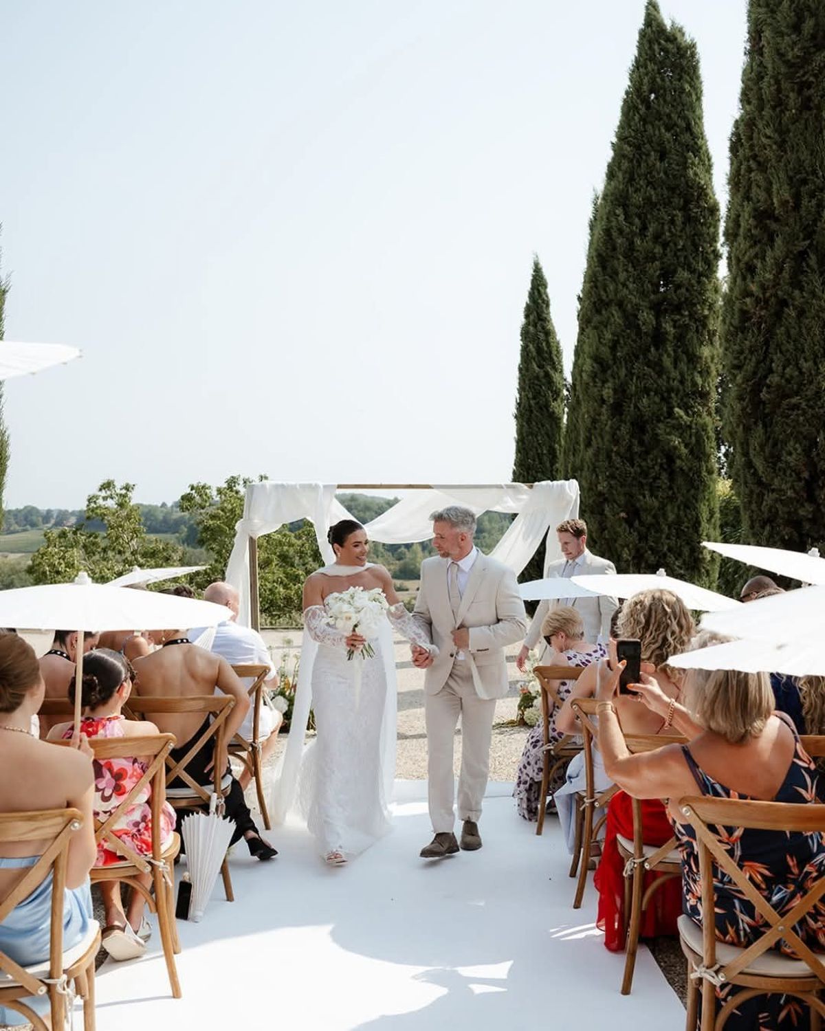 Bride walks the white aisle hand in hand with a grey-haired man in a cream suit, away from a draped arch with hills behind