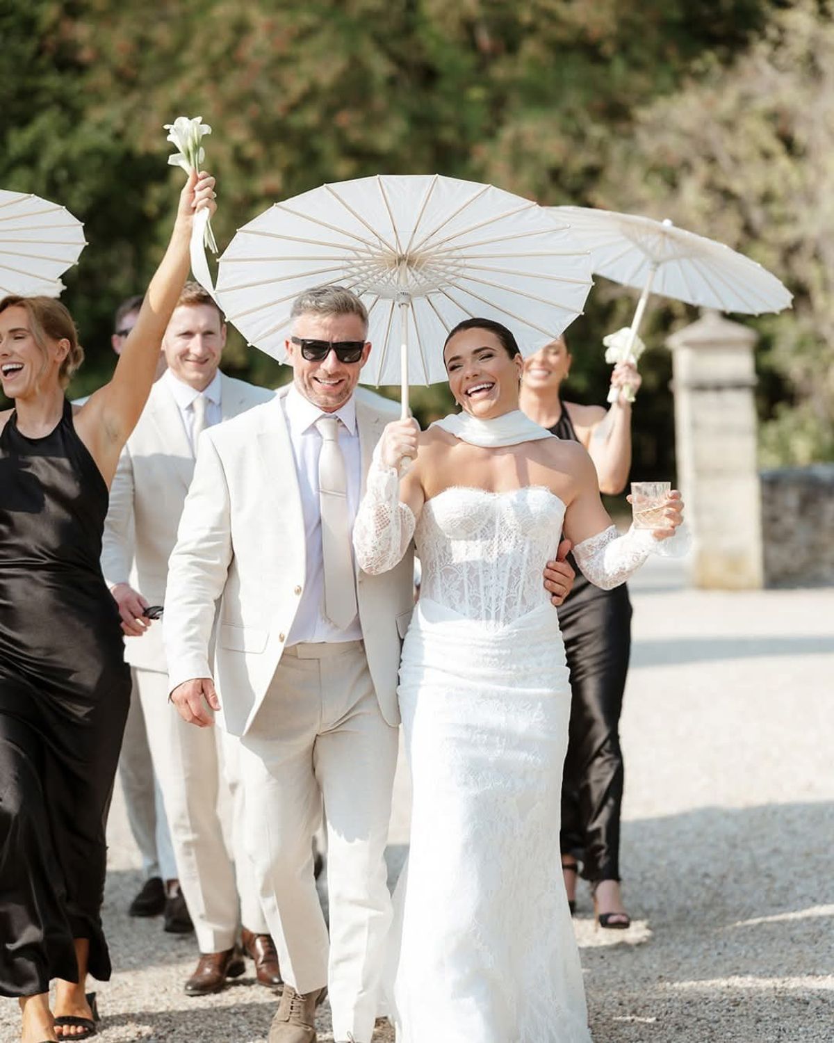 Bride laughs under a white parasol beside a man in a cream suit and sunglasses, guests following on gravel