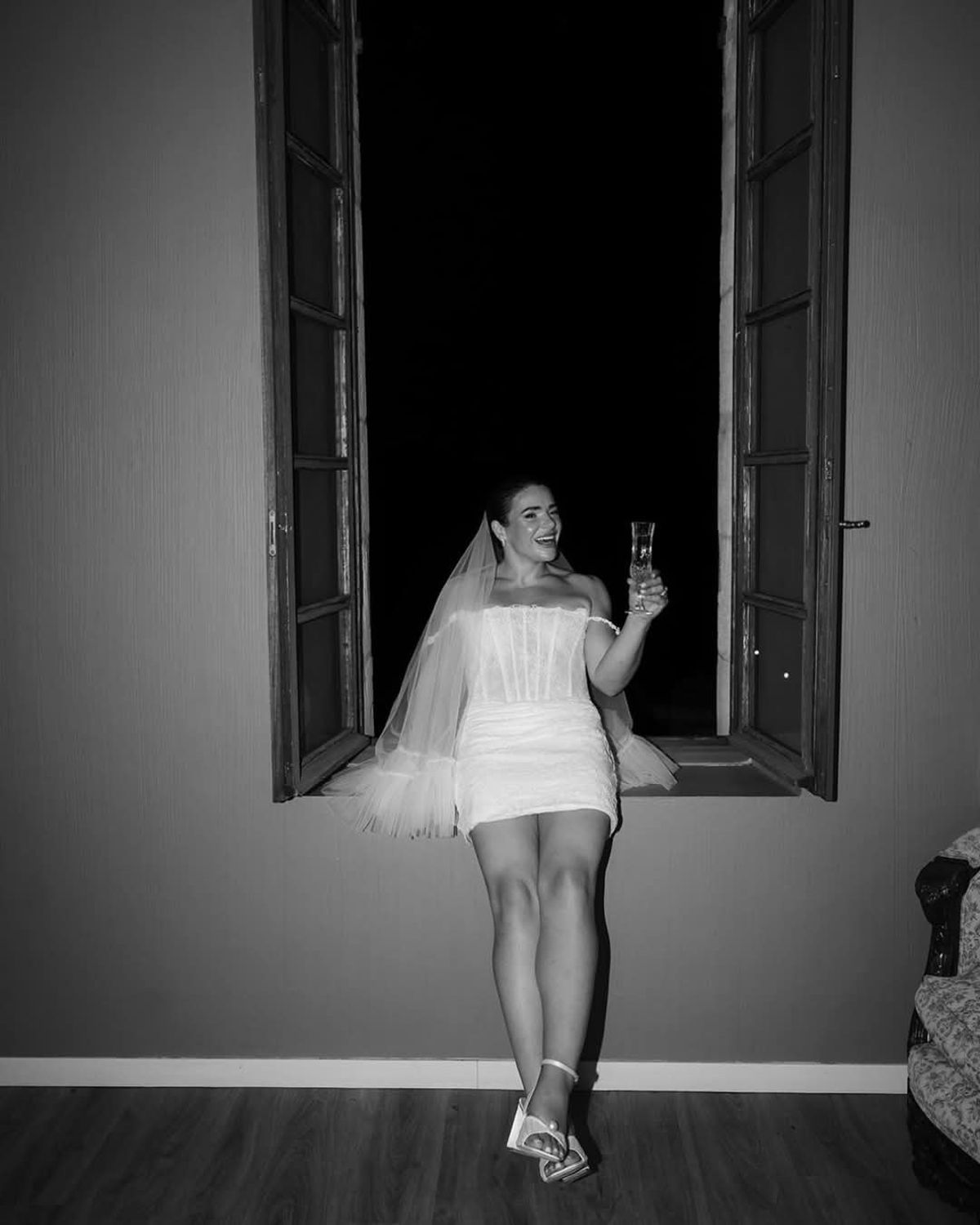 Bride in a short dress and veil sits on an open window sill at night holding a glass, in black and white