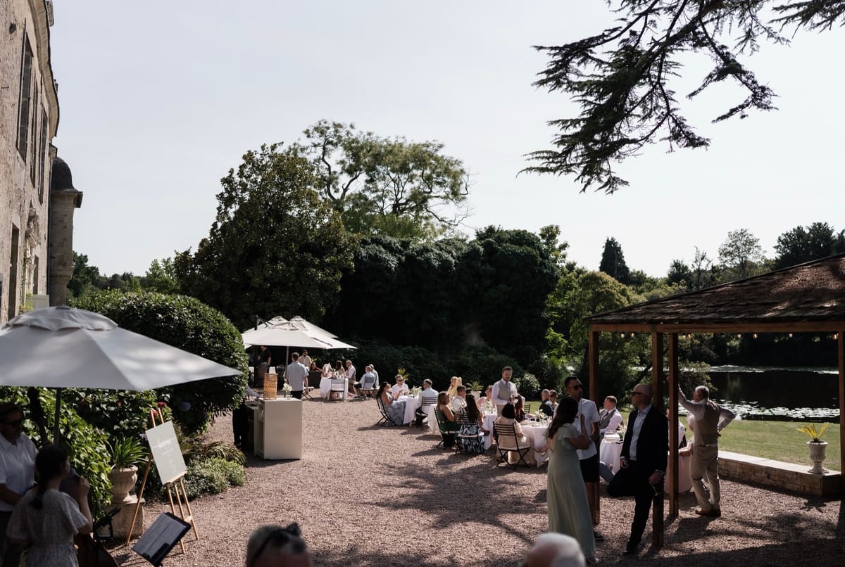 Drinks reception on the gravel terrace at Chateau du Pordor, with parasols and a pergola beside the lake
