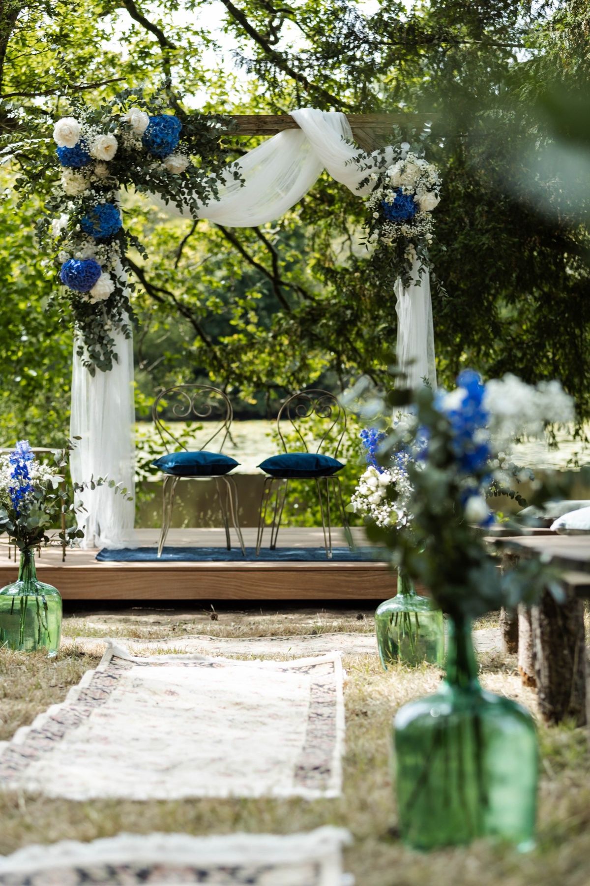 Outdoor ceremony arch at Chateau du Pordor dressed with blue and white flowers, with an aisle runner and green demijohns