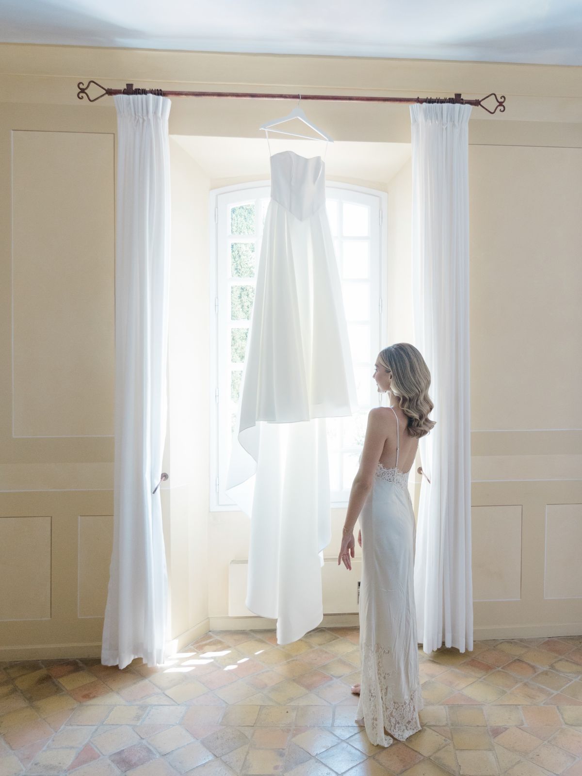 A bride looking at her wedding dress hanging in the window of a chateau guest room