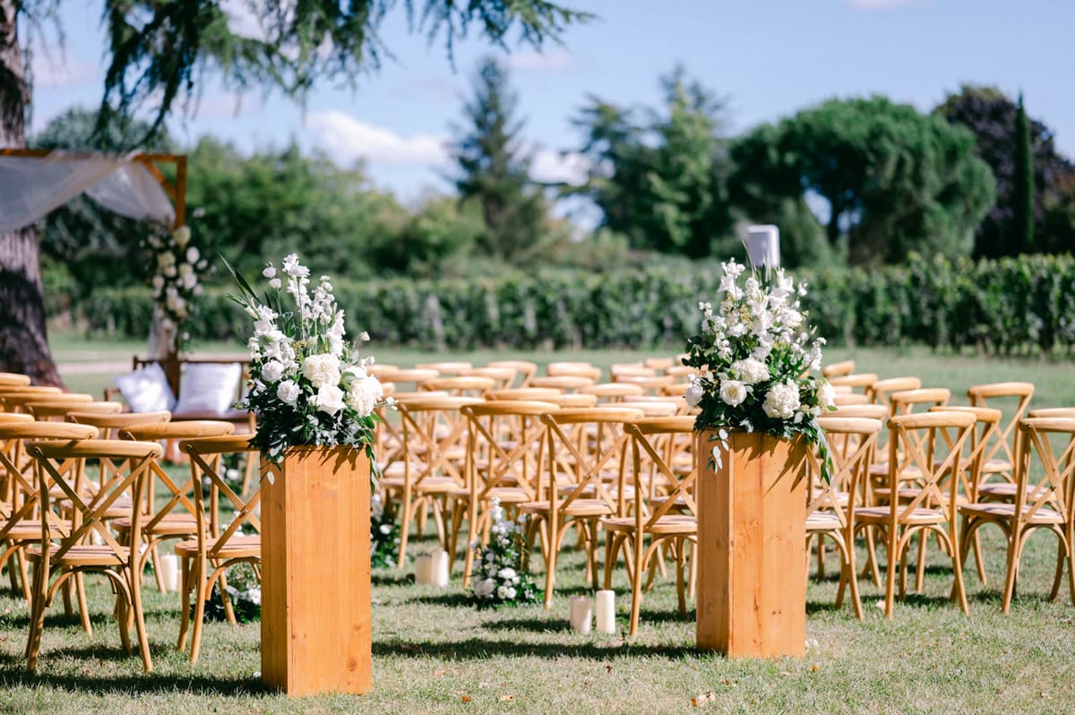Wider view of the outdoor ceremony aisle with cross-back chairs, floral pedestals and candles