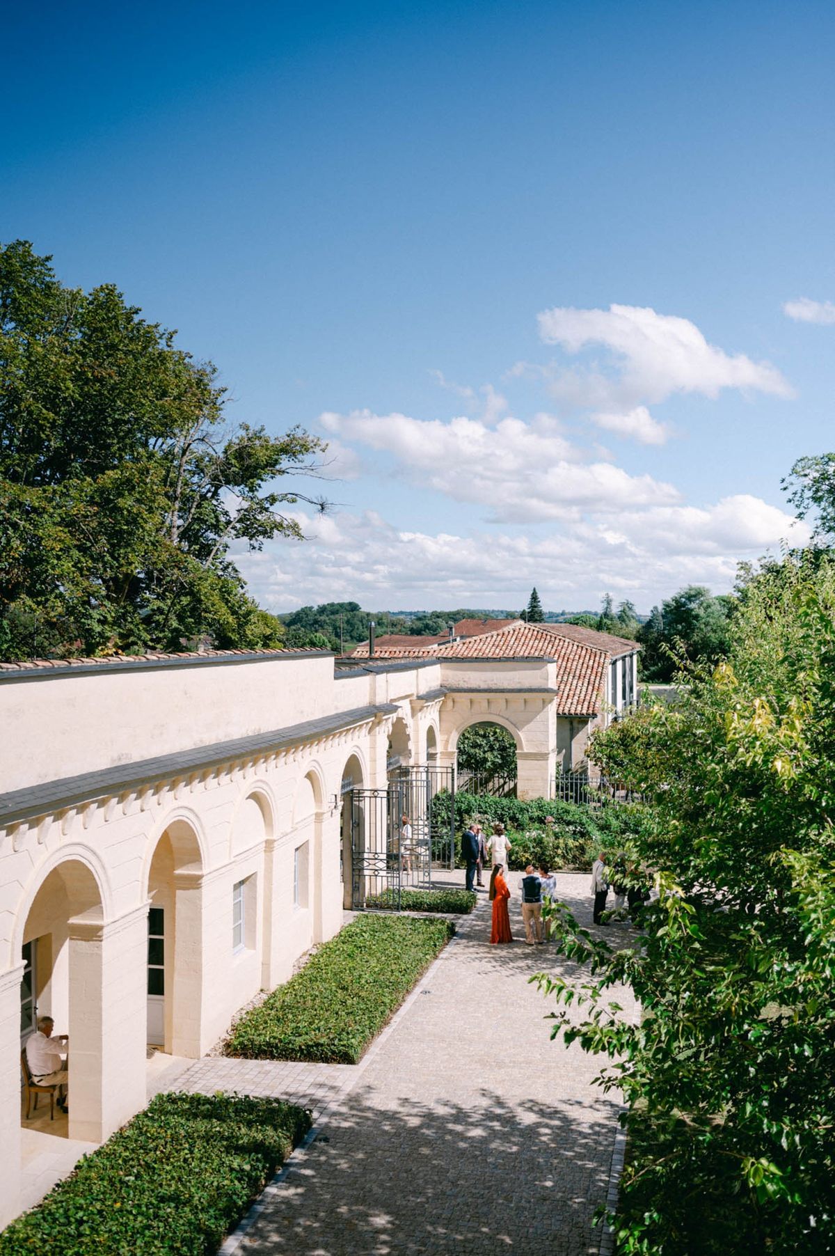 Side view of the arcaded orangery and walled garden at Chateau de l'Hospital, a guest in orange walking the path