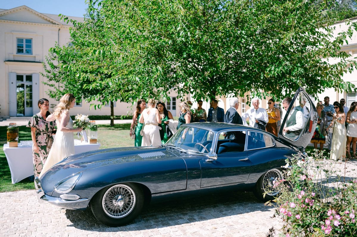 Vintage Jaguar E-Type parked outside Chateau de l'Hospital with the couple and guests gathered around