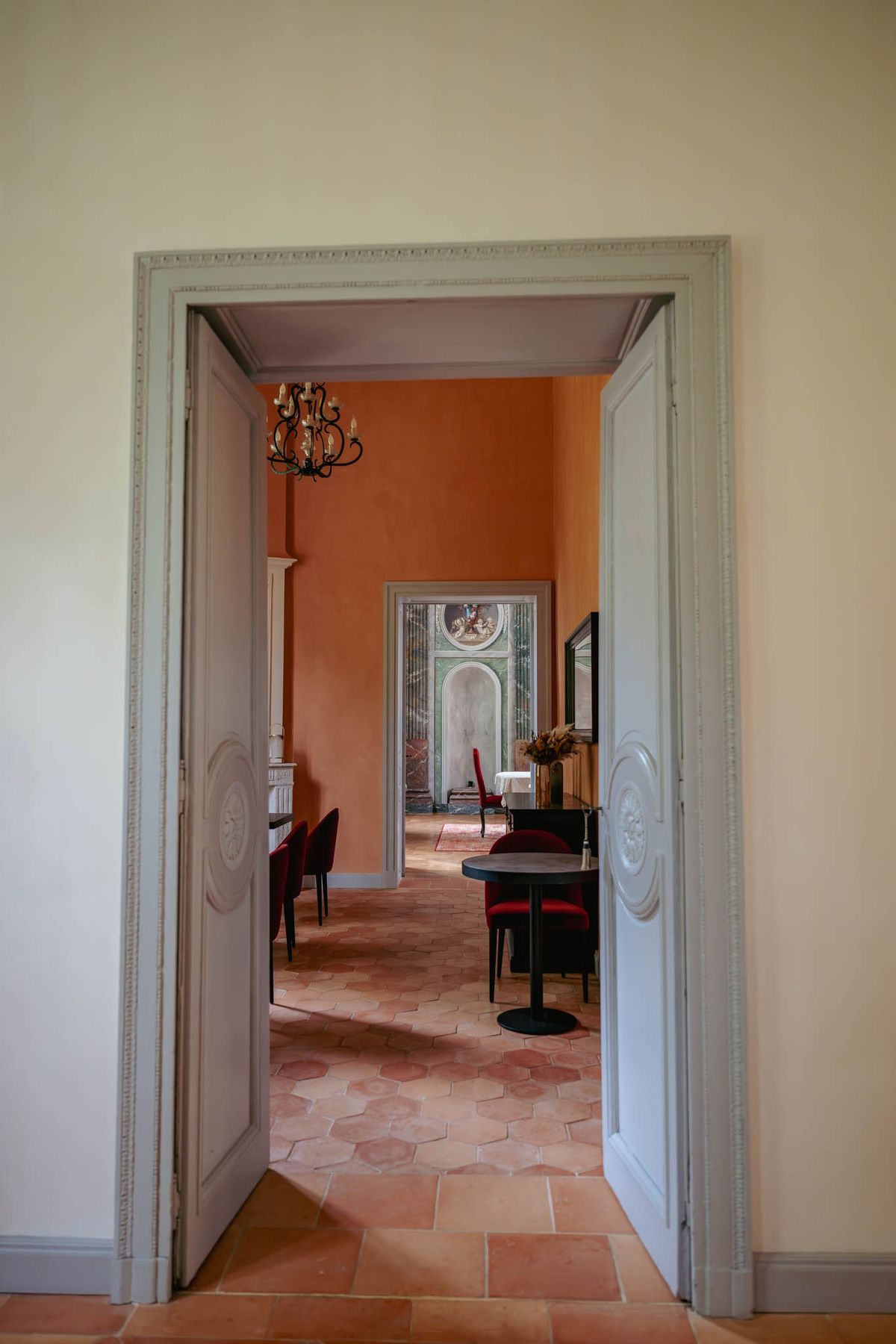 Enfilade view through the Dining Room at Chateau de l'Hospital: terracotta hexagon floor, orange walls, chandelier