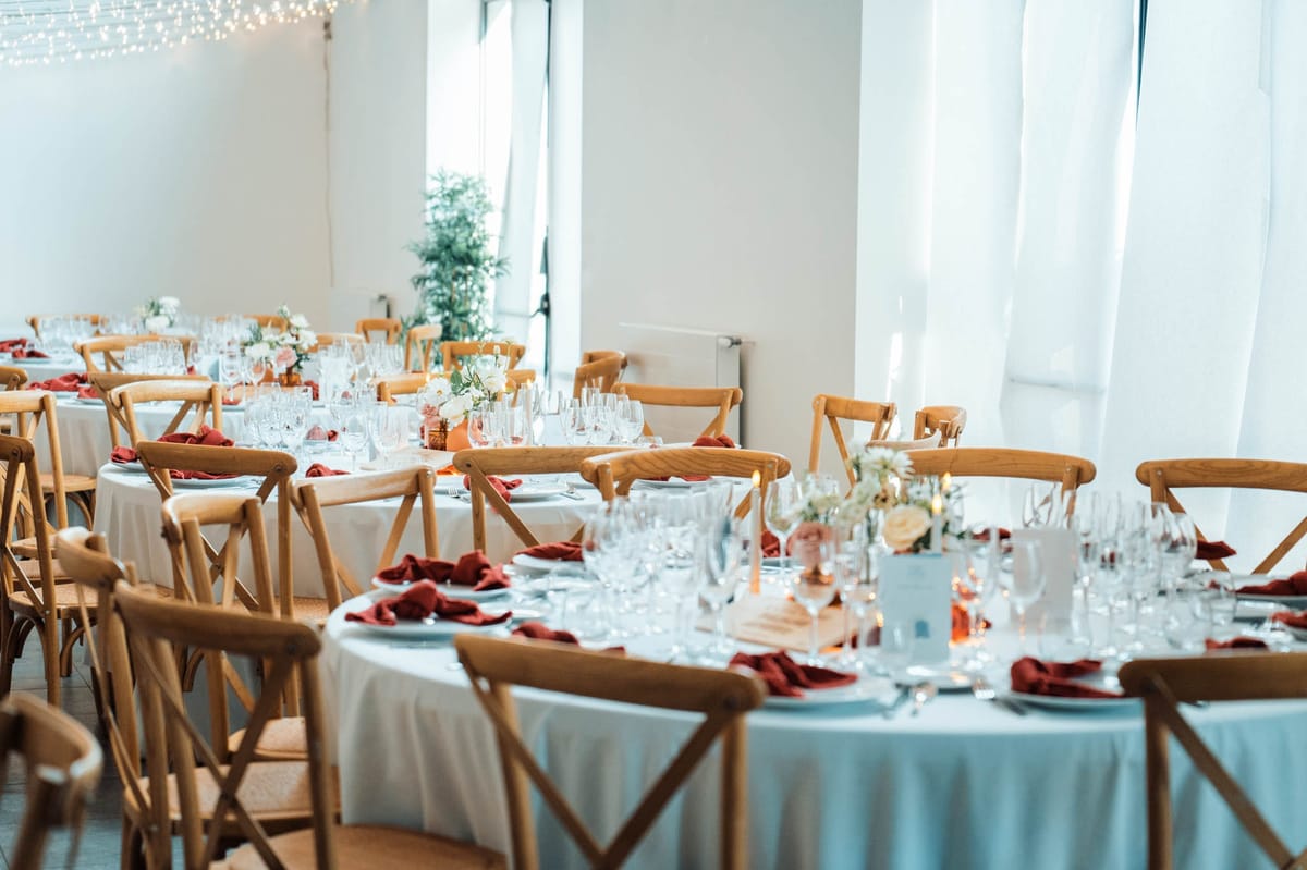 Orangery Hall at Chateau de l'Hospital set for a seated wedding dinner with round tables, red napkins and fairy-light canopy