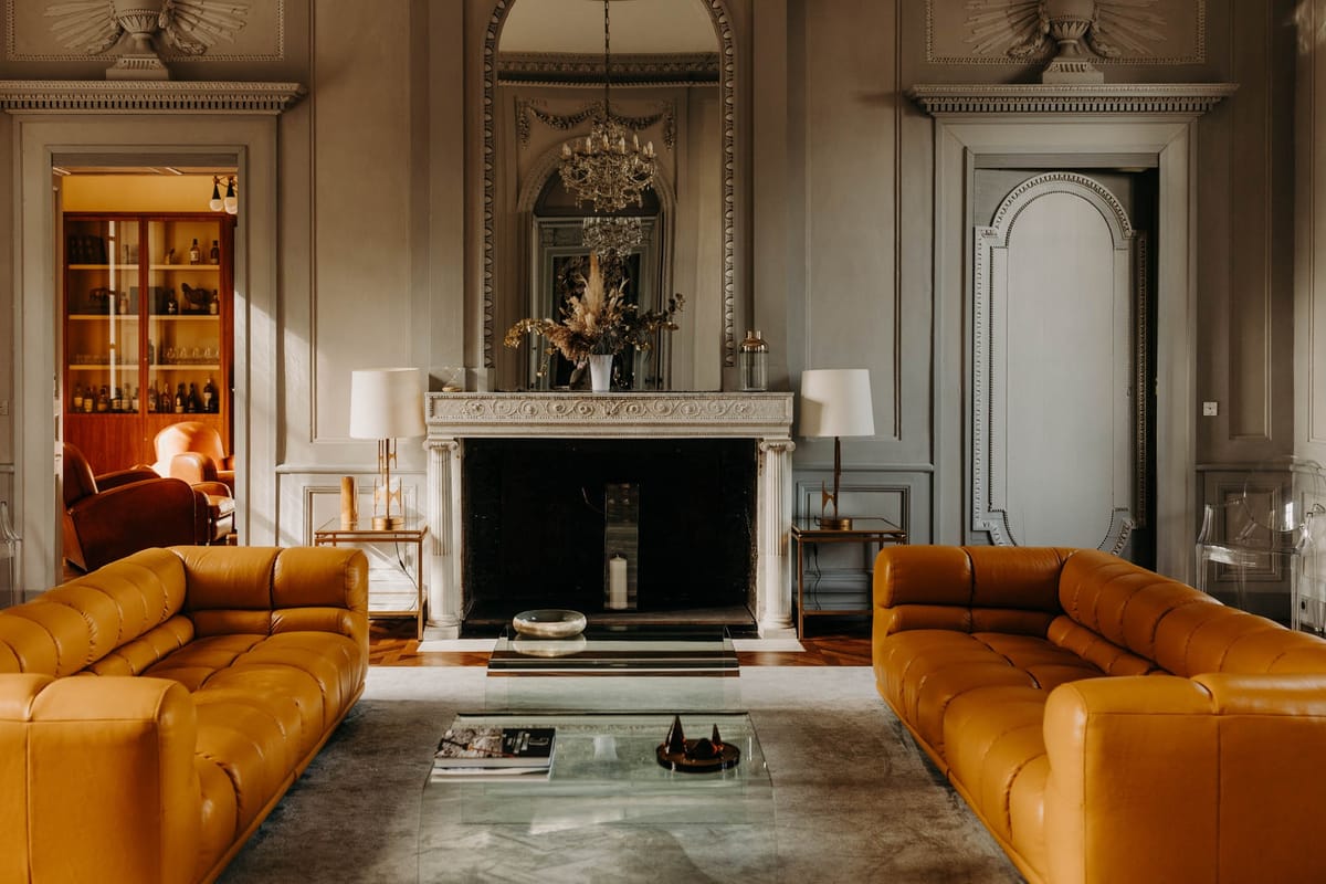 Grand salon at Chateau de l'Hospital with panelled grey walls, caramel leather sofas facing a marble fireplace and chandelier above