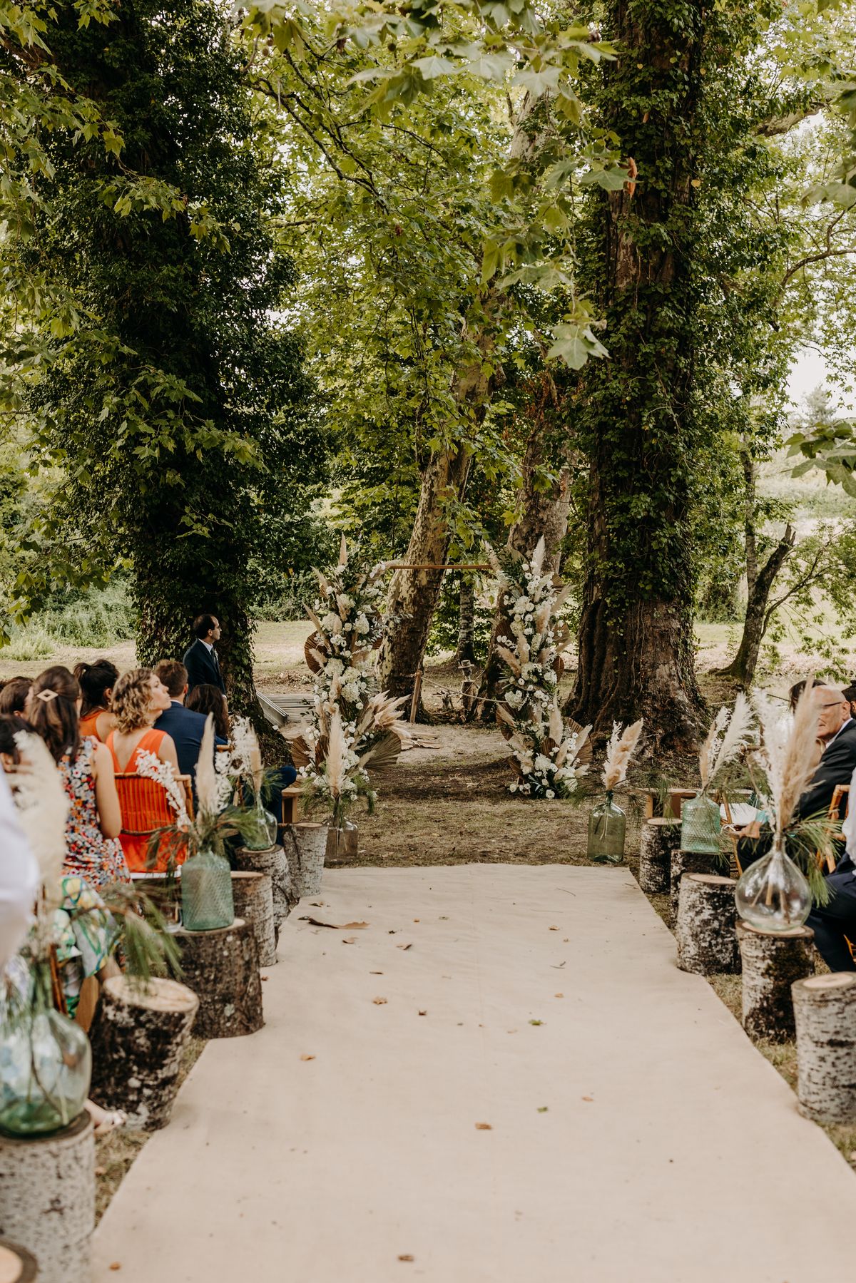 Outdoor wedding ceremony aisle set beneath mature trees beside the estate's water, with bamboo chairs and dried-flower arrangements