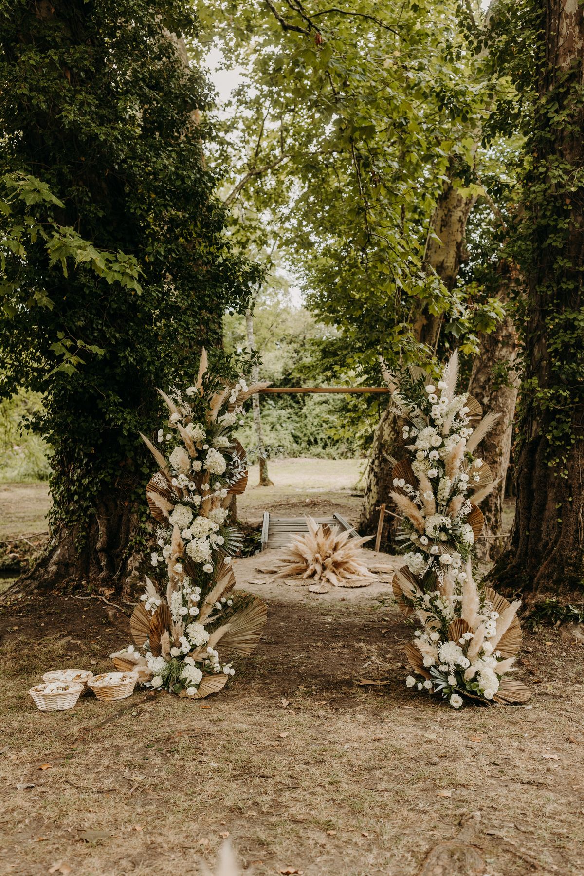 A dried pampas grass and white floral ceremony arch framed by an avenue of mature trees