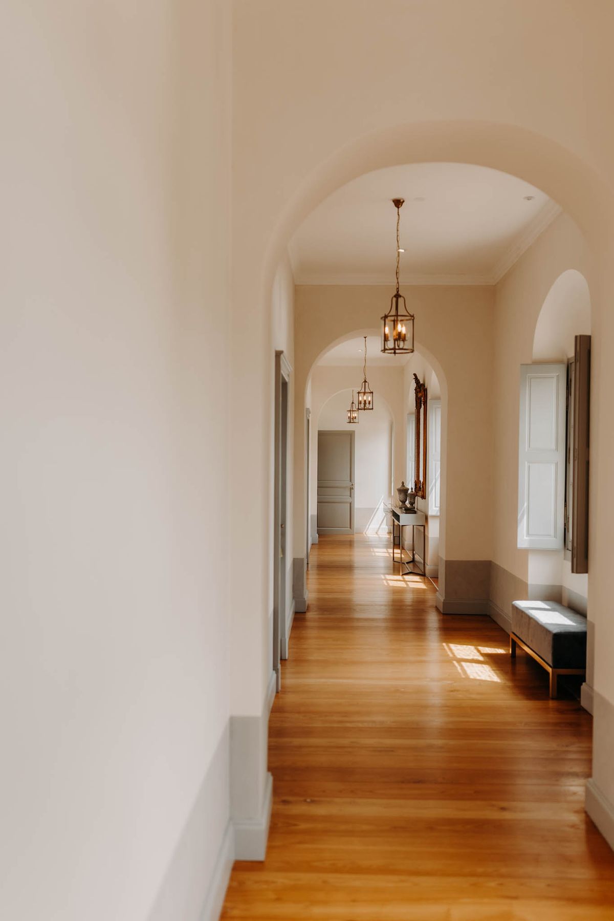 Sunlit arched corridor in Château de l'Hospital with brass lantern pendants and wooden floors