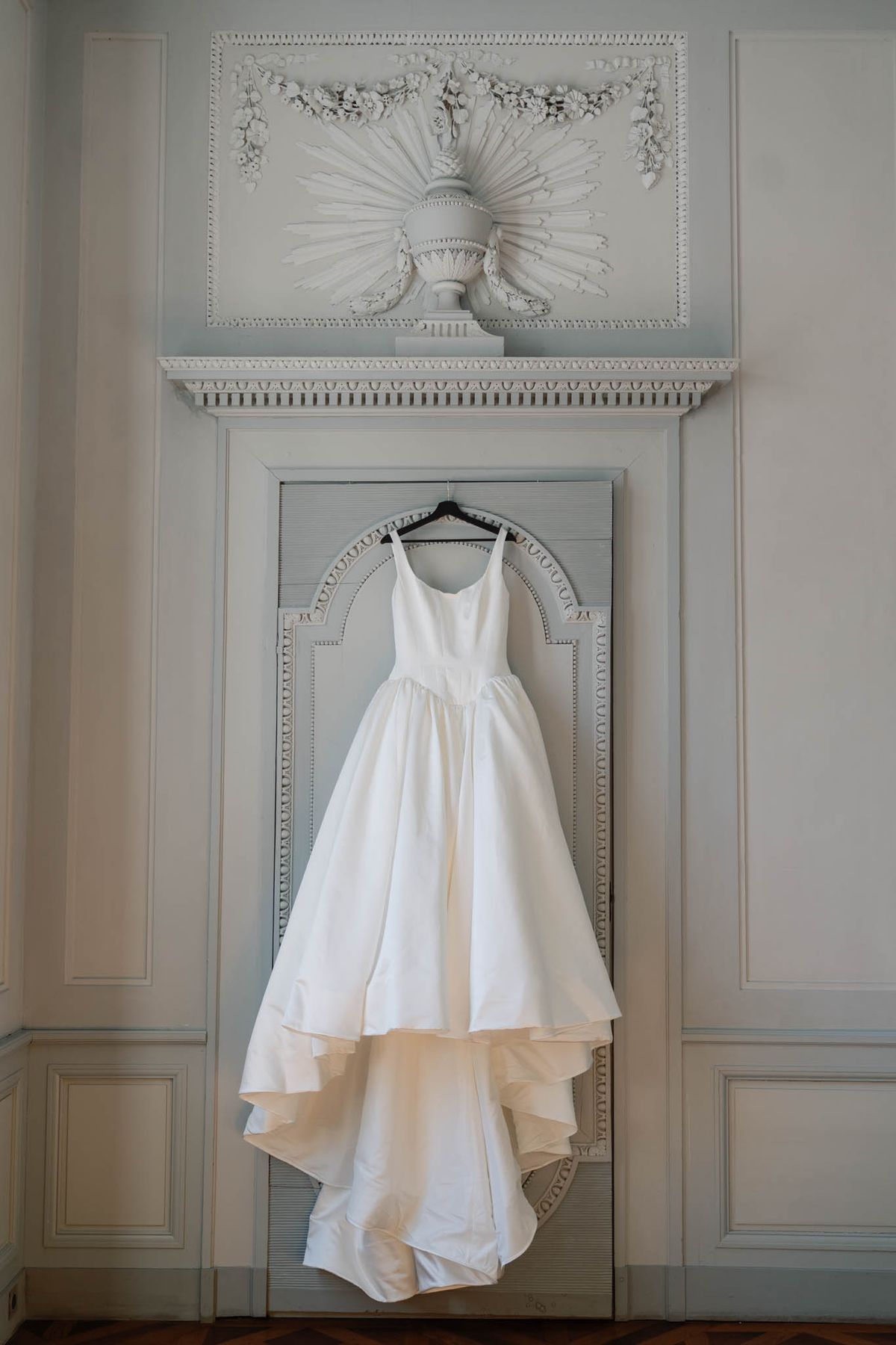 Ivory wedding gown hanging against a panelled door beneath a sculpted plaster urn motif