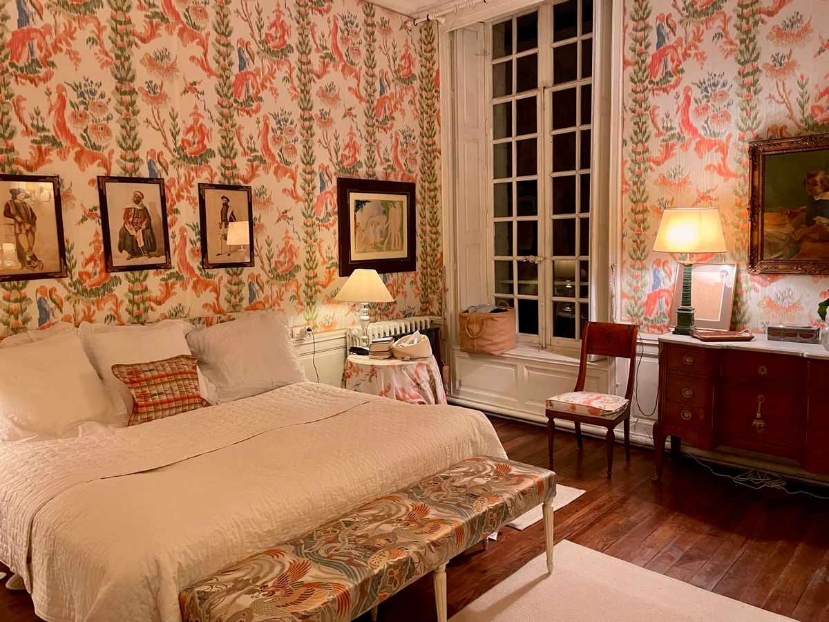 A guest bedroom papered in coral toile with tall French windows and a mahogany chest