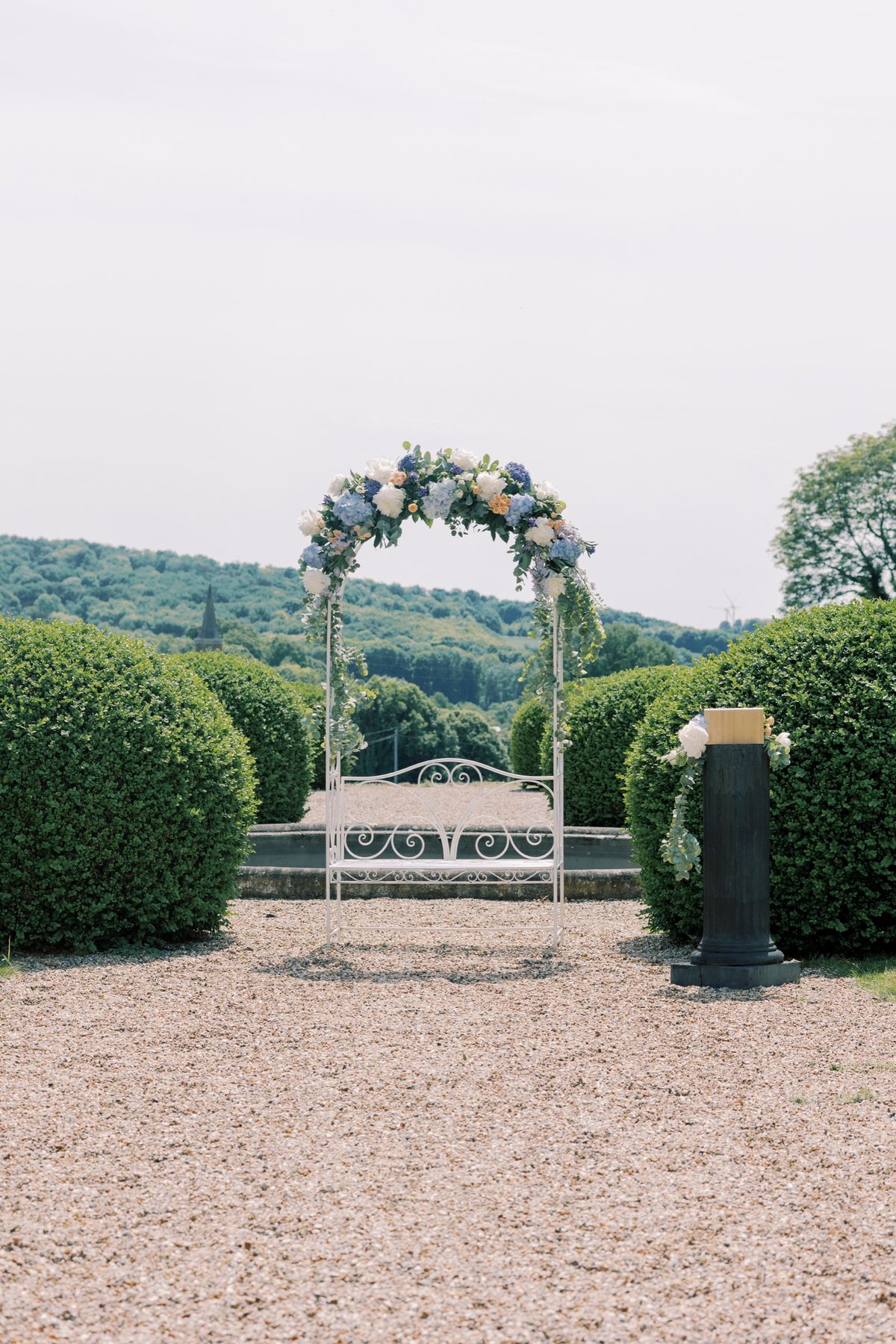 The outdoor ceremony arch dressed with flowers and set with a bench, framed by topiary and the valley view
