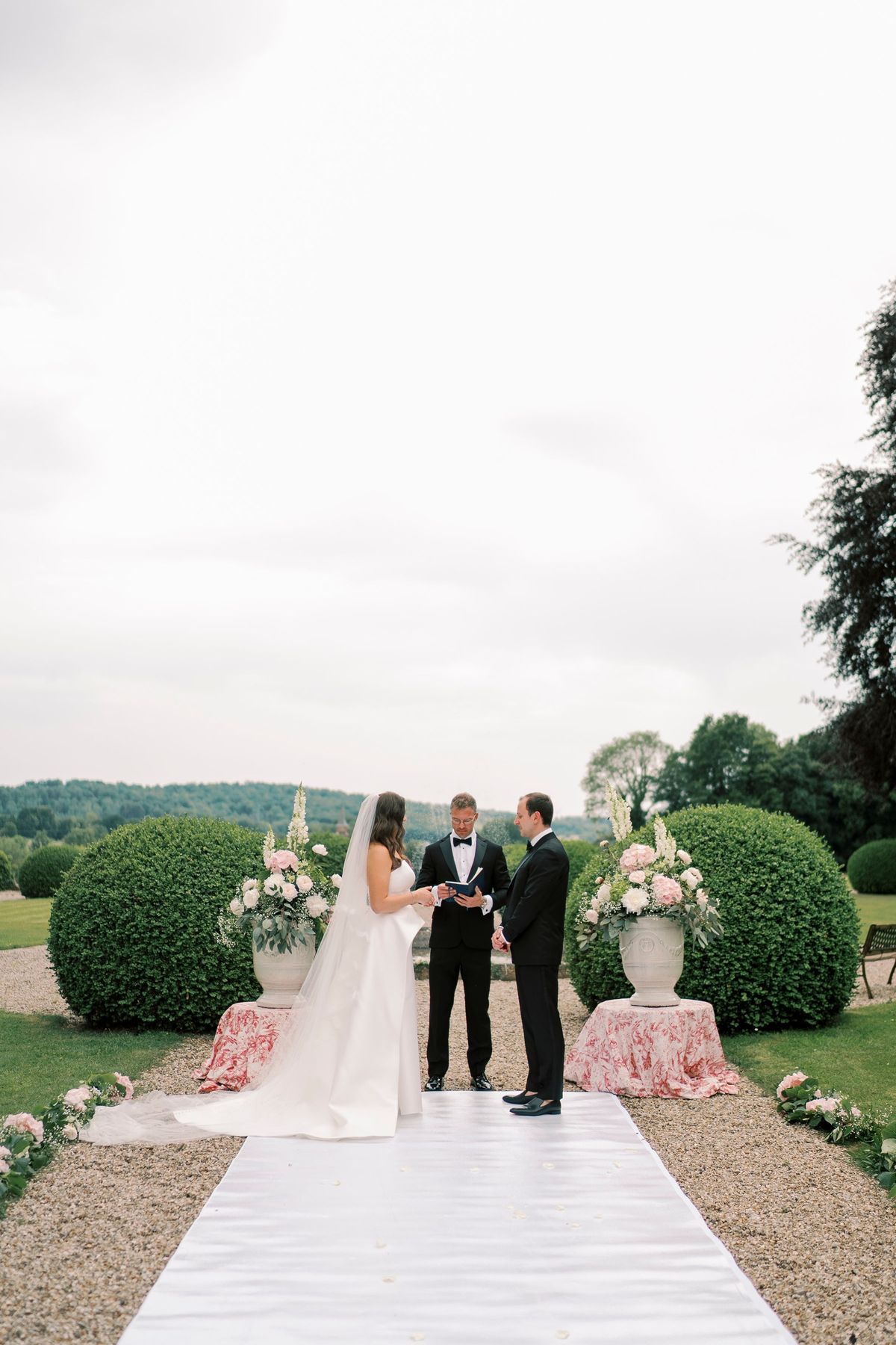 A couple exchanging vows on the garden aisle with urns of pink and white flowers