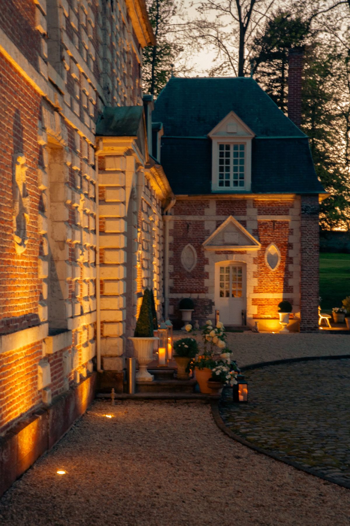 The château and its garden pavilion lit at dusk, with potted flowers on the gravel